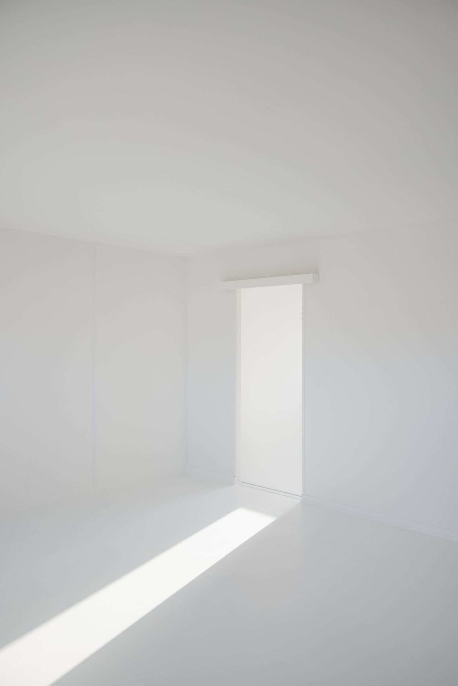 Empty all-white room with bright natural light streaming through a doorway casting shadows on floor.