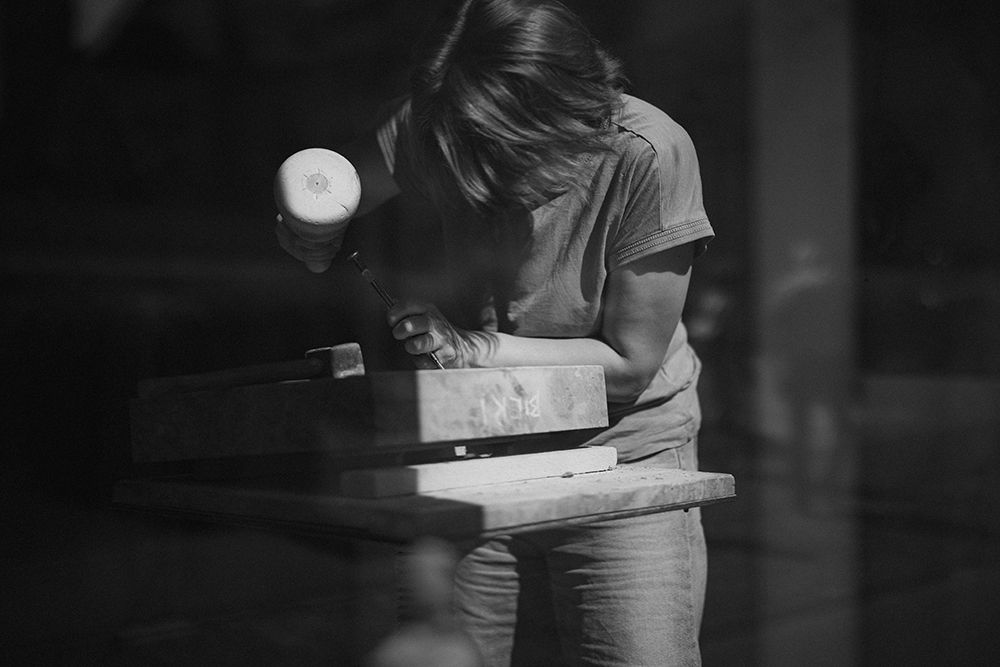 Black and white photo of a sculptor carefully carving stone with a mallet in a dark studio.