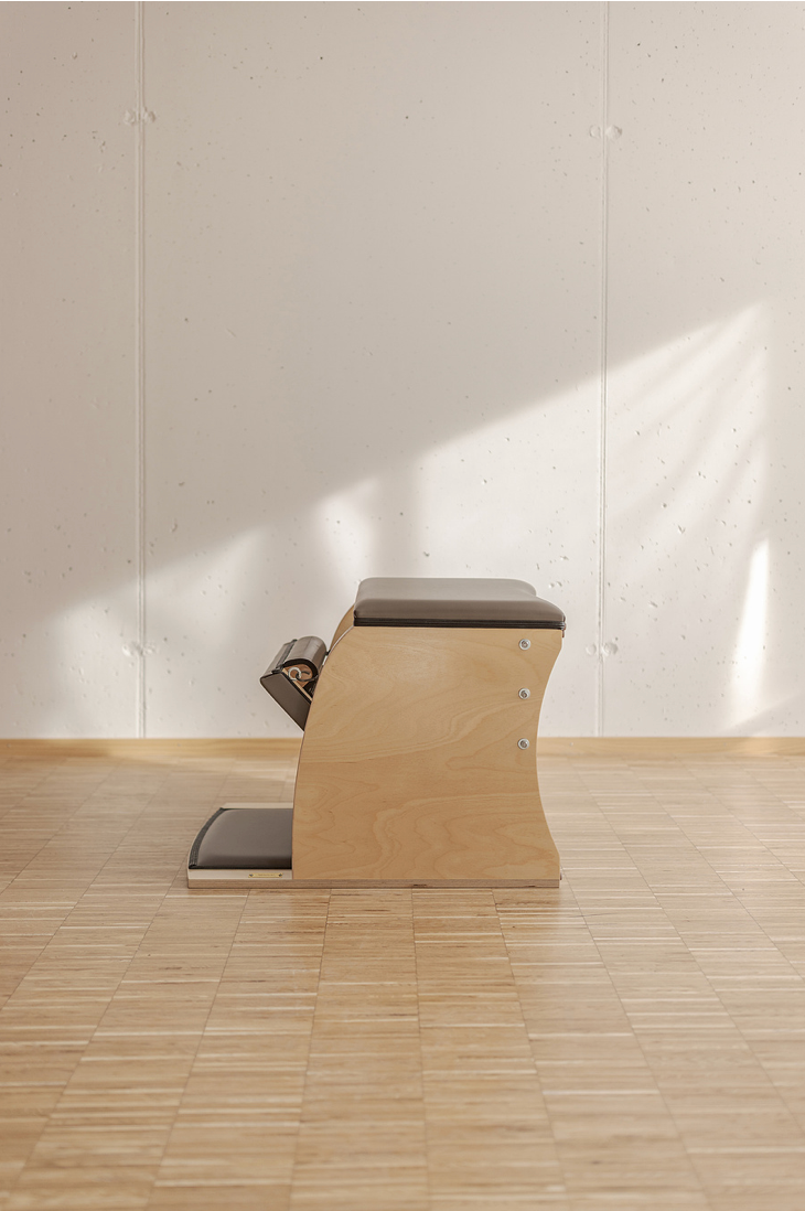Wooden Pilates wunda chair with dark padding on light hardwood floor against white wall.