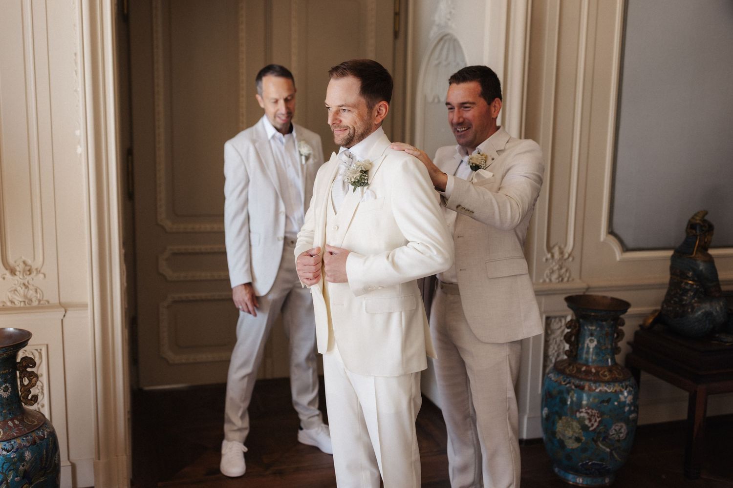 Groom in white suit being helped by groomsmen in elegant room before wedding ceremony.