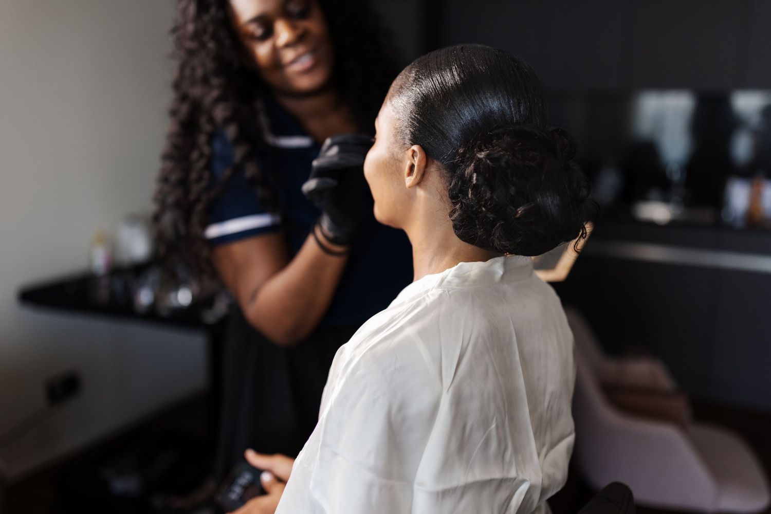 Makeup artist in black gloves styling a bride with an elegant updo in a salon setting.