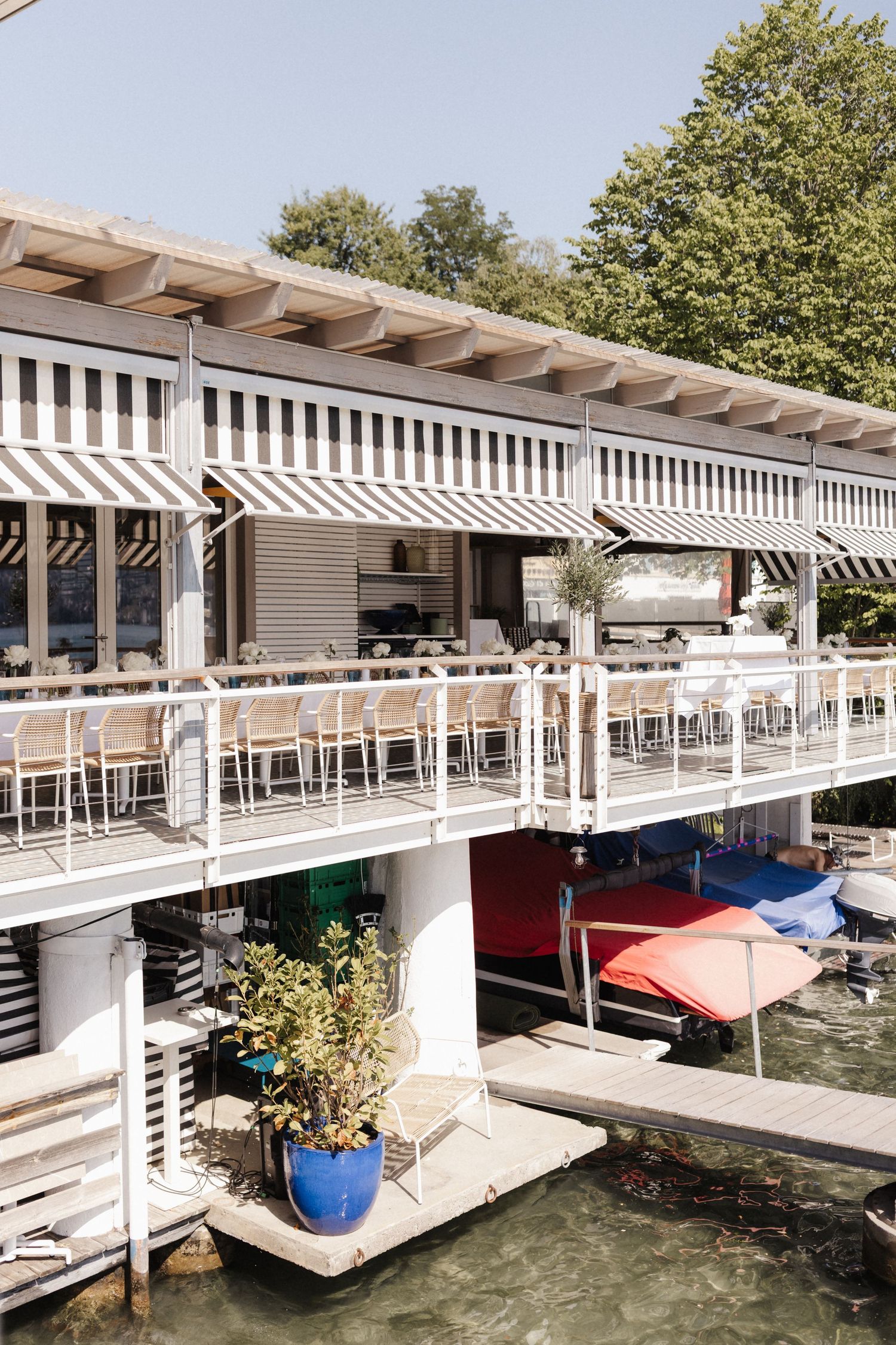 Two-story waterfront restaurant with striped awnings, white railings, and boats docked below.