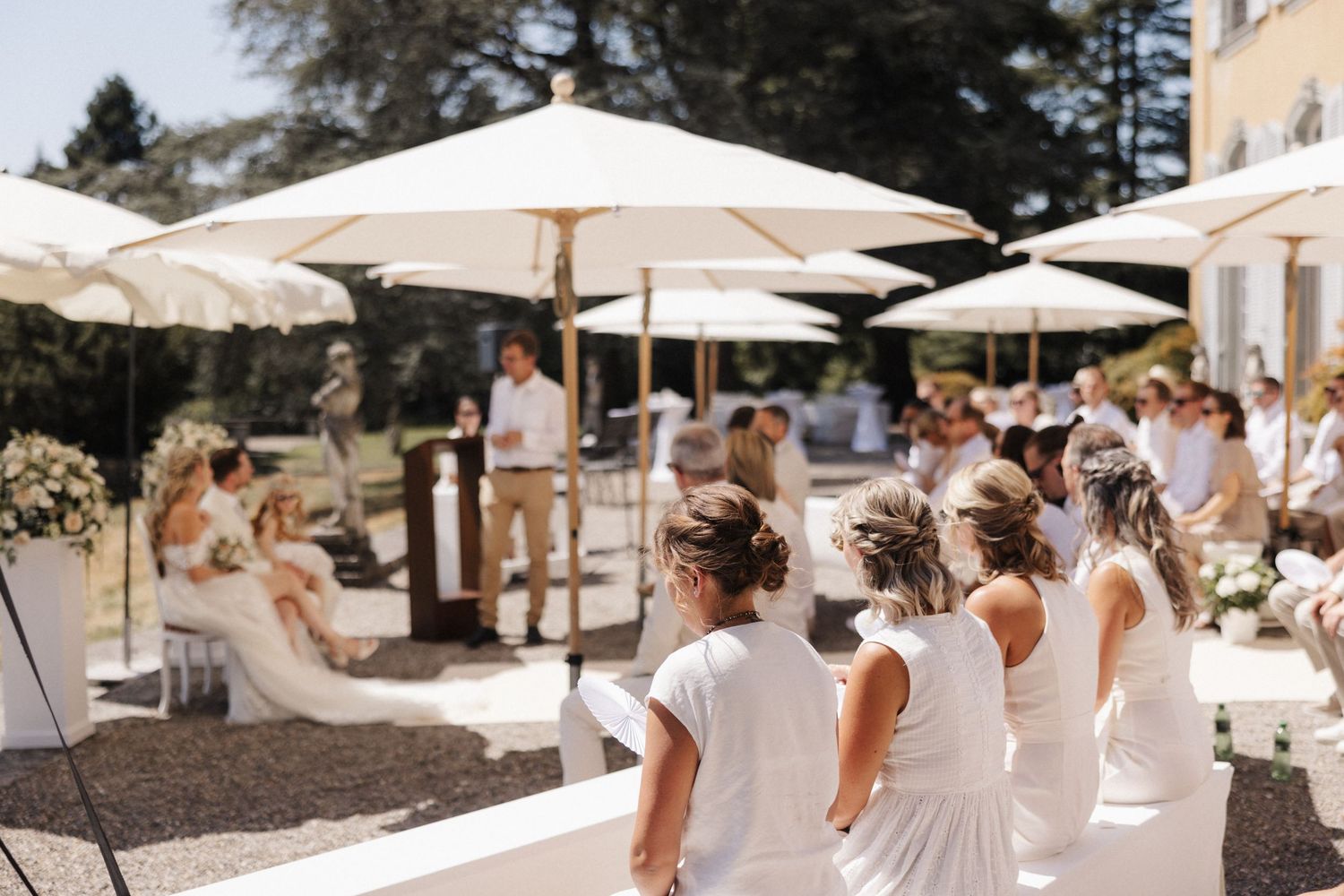 Bridesmaids in white dresses watch an outdoor wedding ceremony under large white umbrellas on a sunny day.