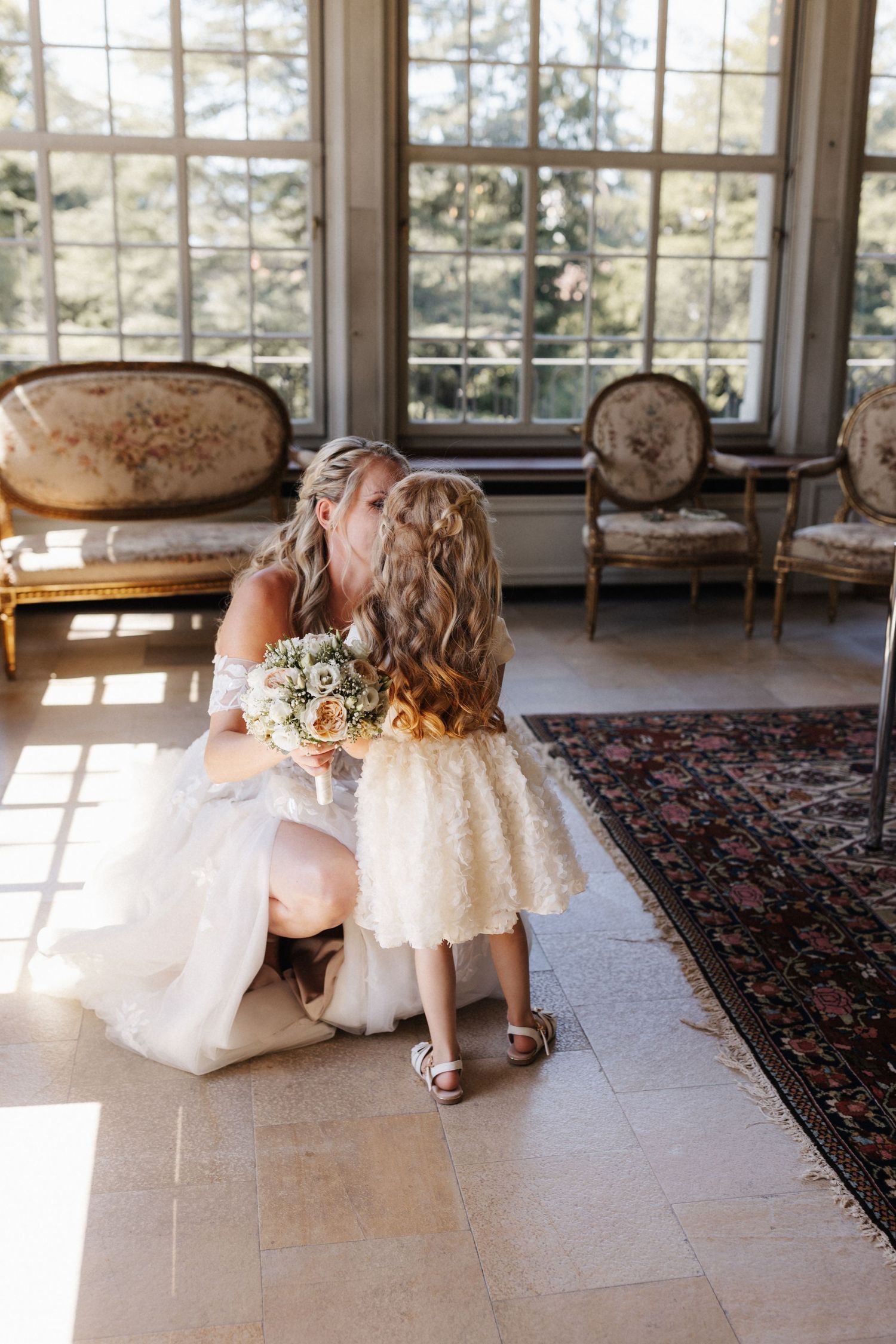 Bride in a white lace gown kneels to kiss a young flower girl in a ruffled white dress indoors.