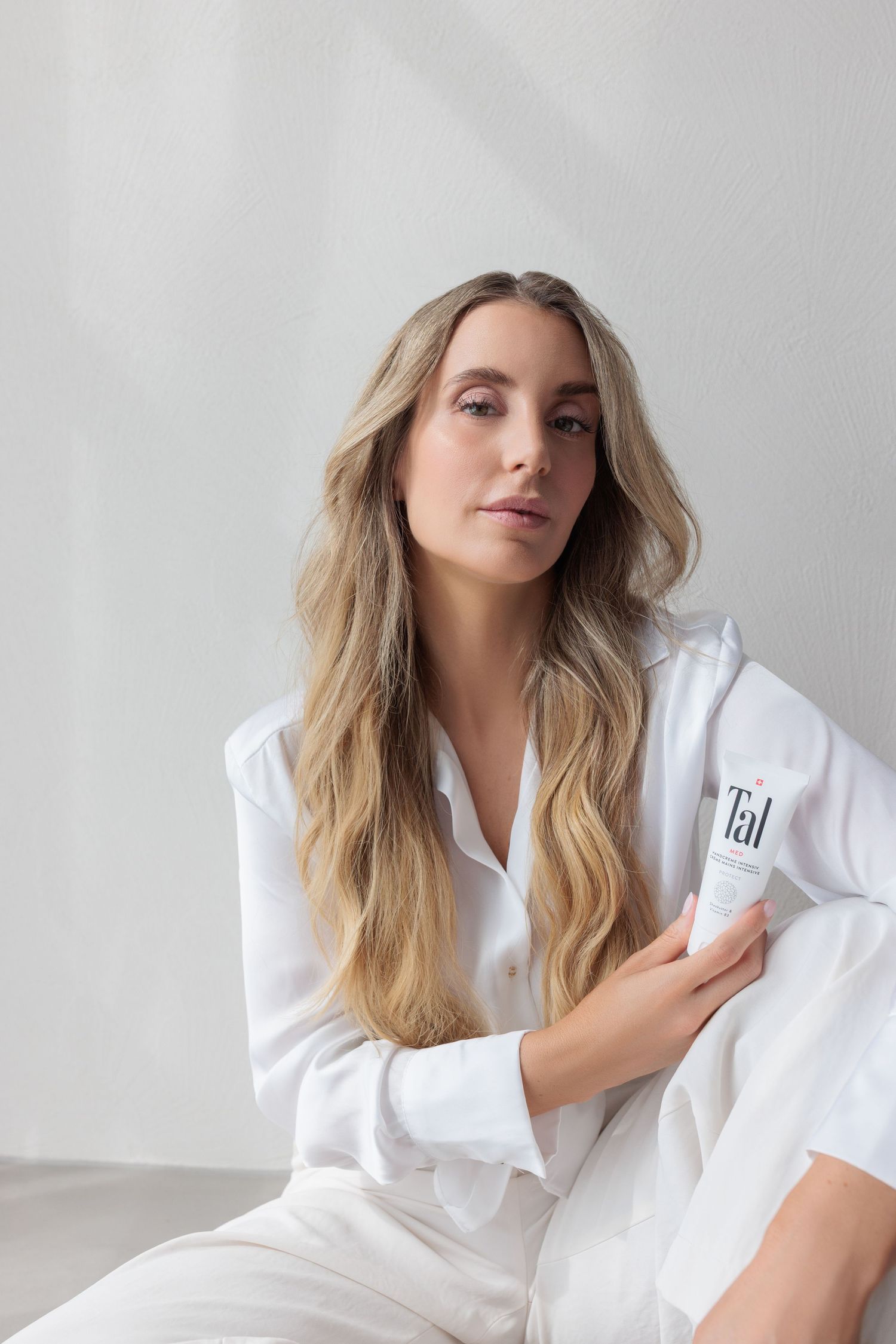 Blonde woman in white shirt sitting and holding Tal skincare tube in bright minimalist room.