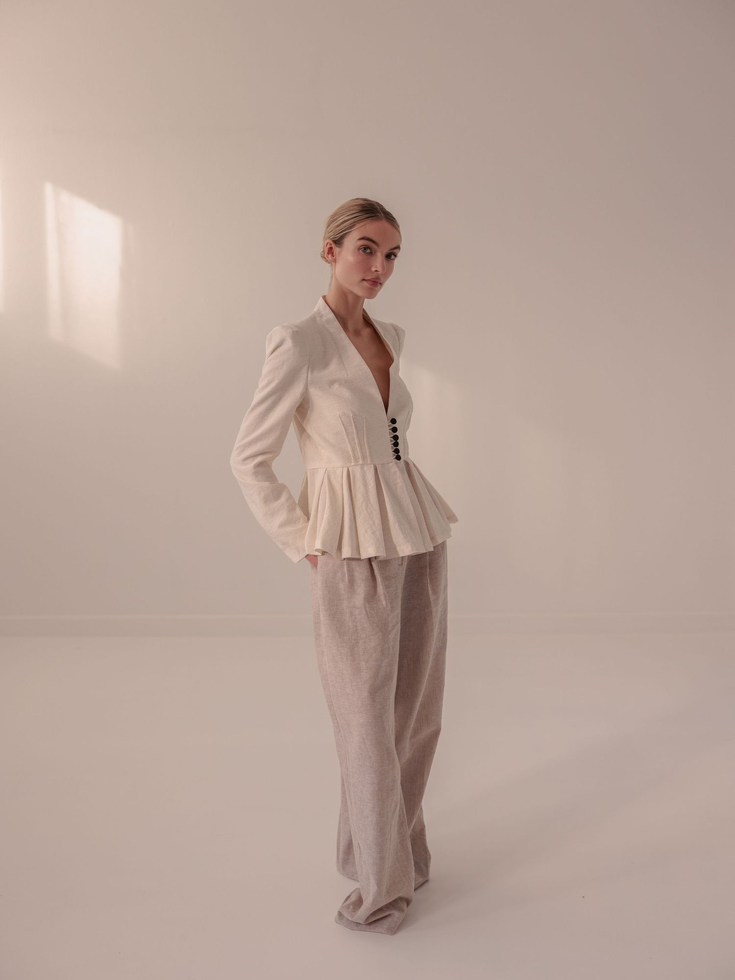 Model in a white structured peplum blazer and wide-leg linen trousers standing in a minimalist studio.