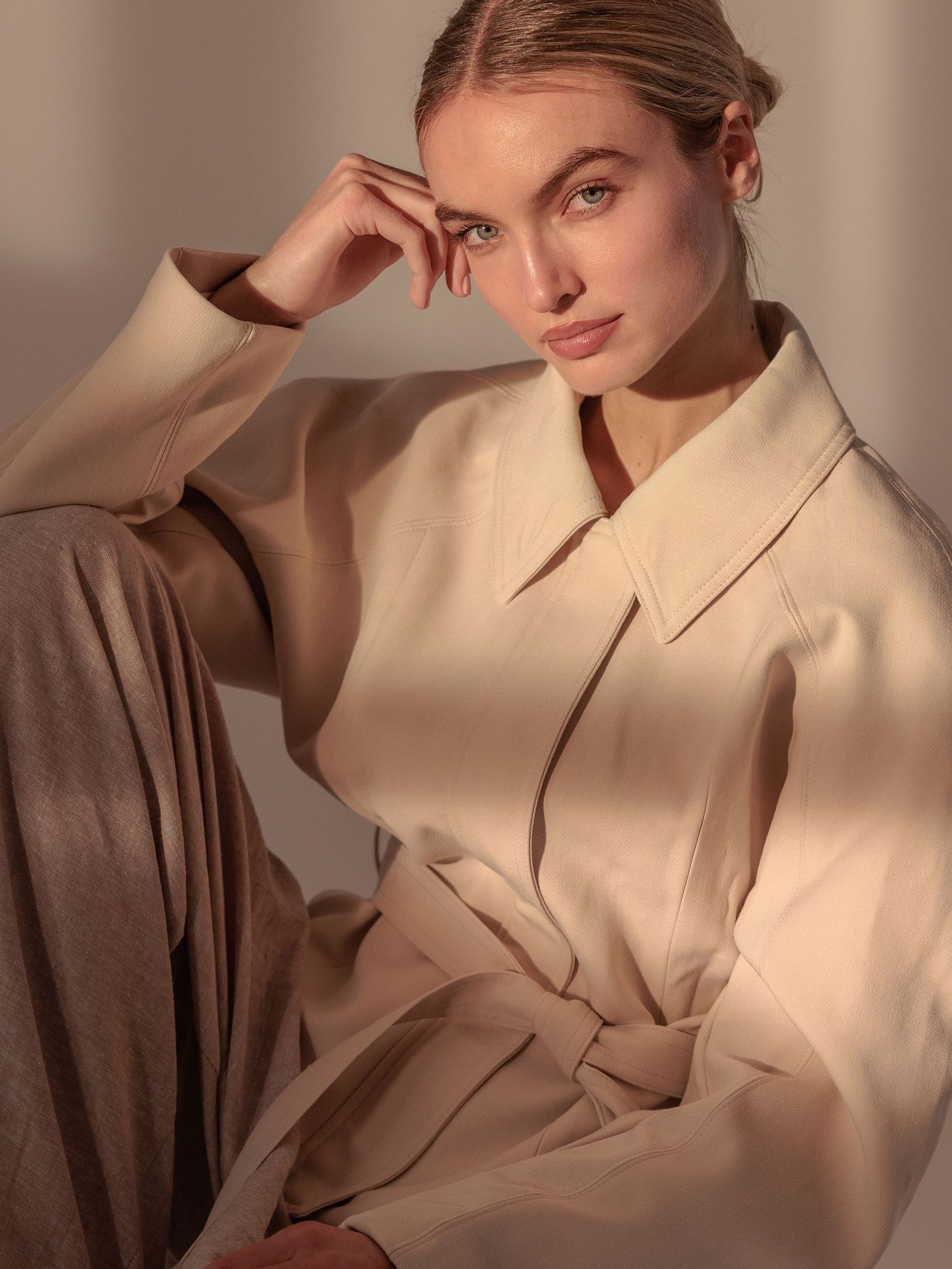 Woman in a cream collared jacket sits with hand to face, bathed in soft warm natural light.