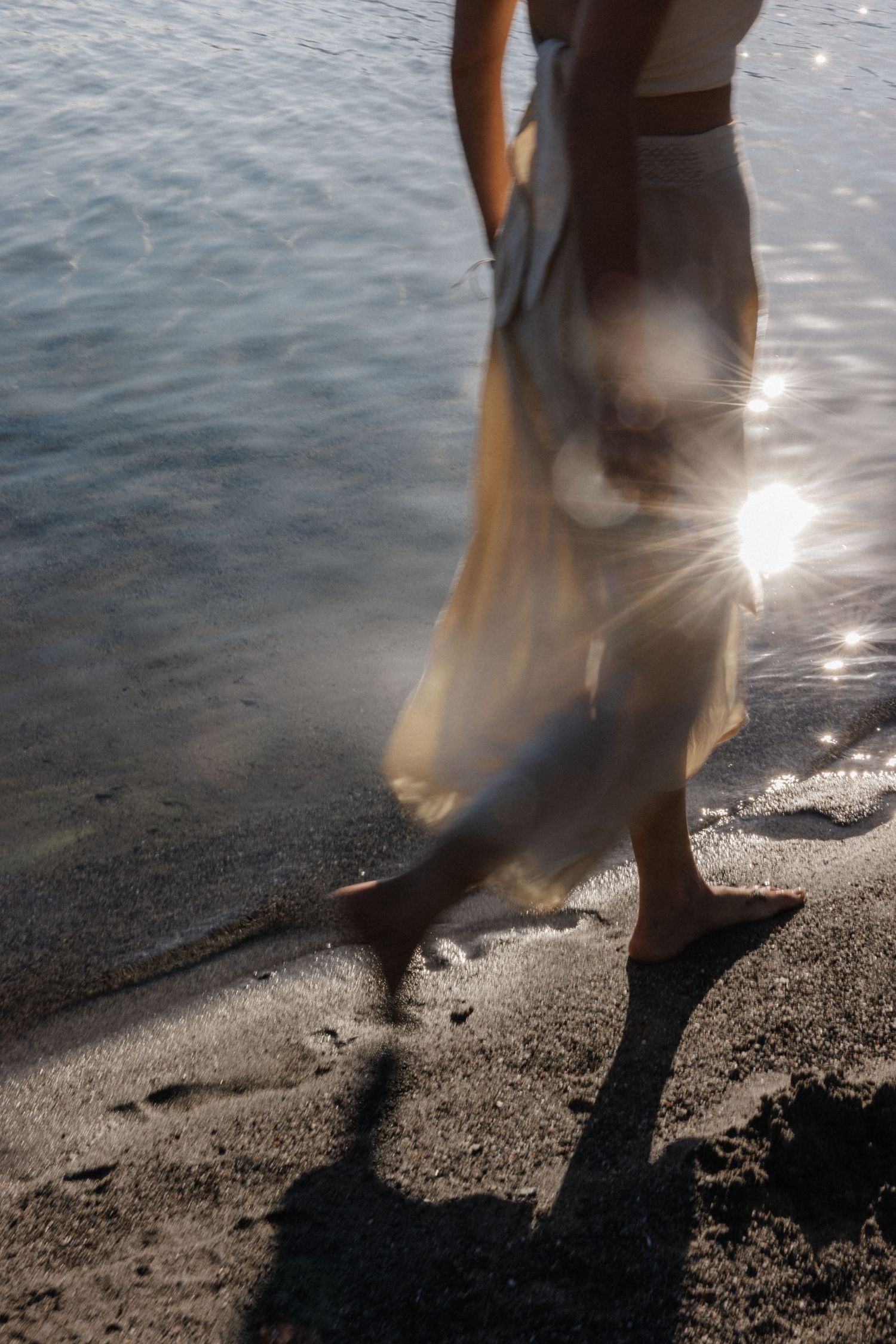 Motion blur of woman in golden dress walking barefoot on sandy beach with sunlight and water behind.