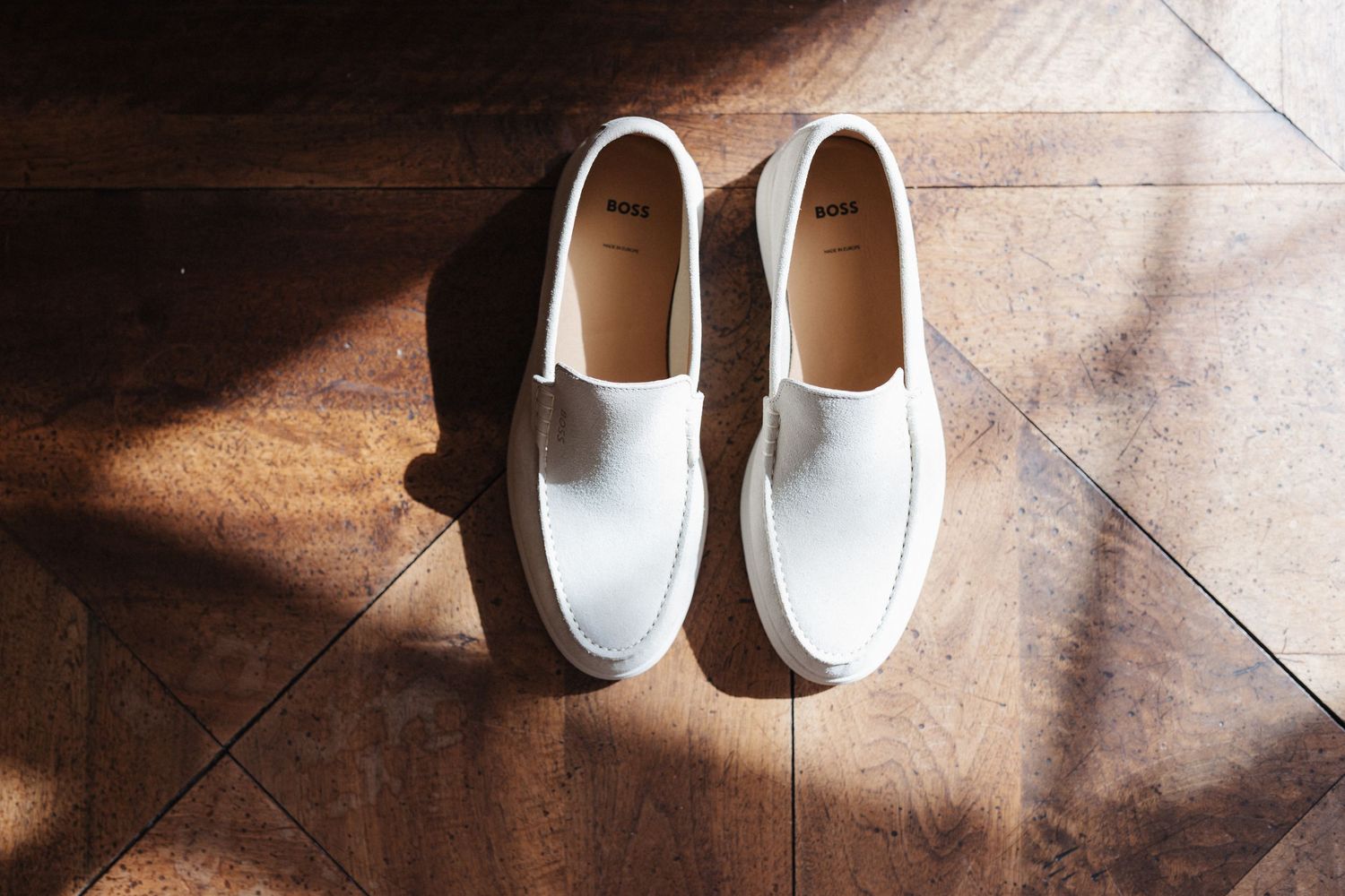 White leather slip-on sneakers with brand label on warm wooden herringbone floor in sunlight.
