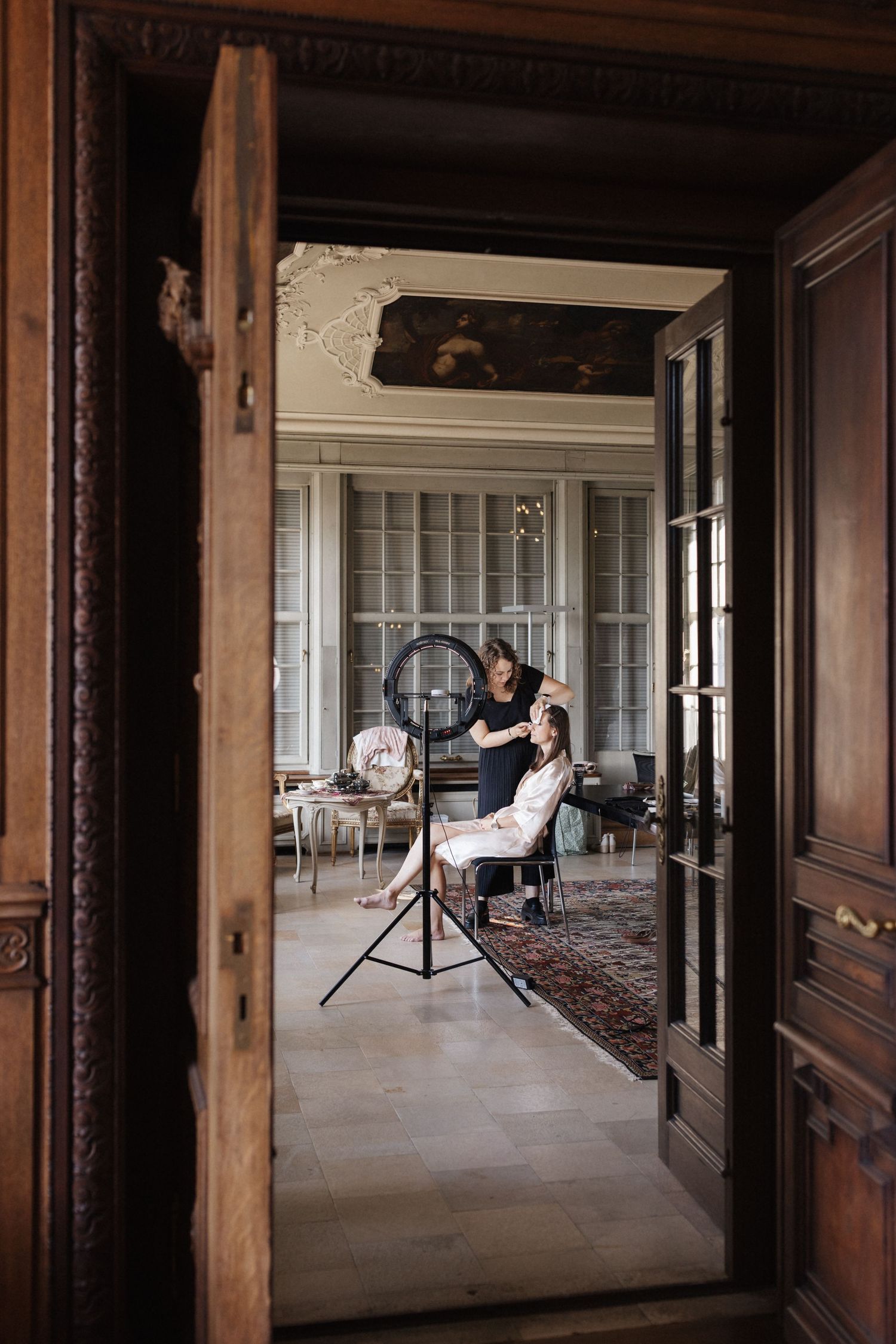 Bride getting hair styled in ornate vintage room viewed through carved wooden doorway.