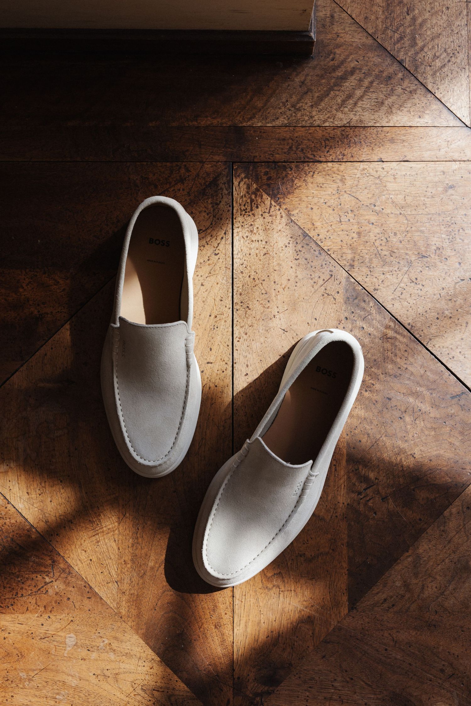 Pair of white suede slip-on loafers on herringbone parquet wooden floor in sunlight.