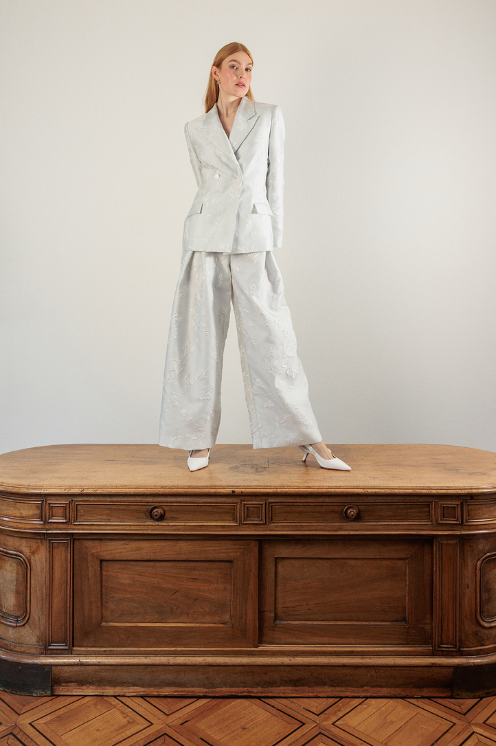 Blonde model in silver linen suit standing on wooden antique desk against white background.