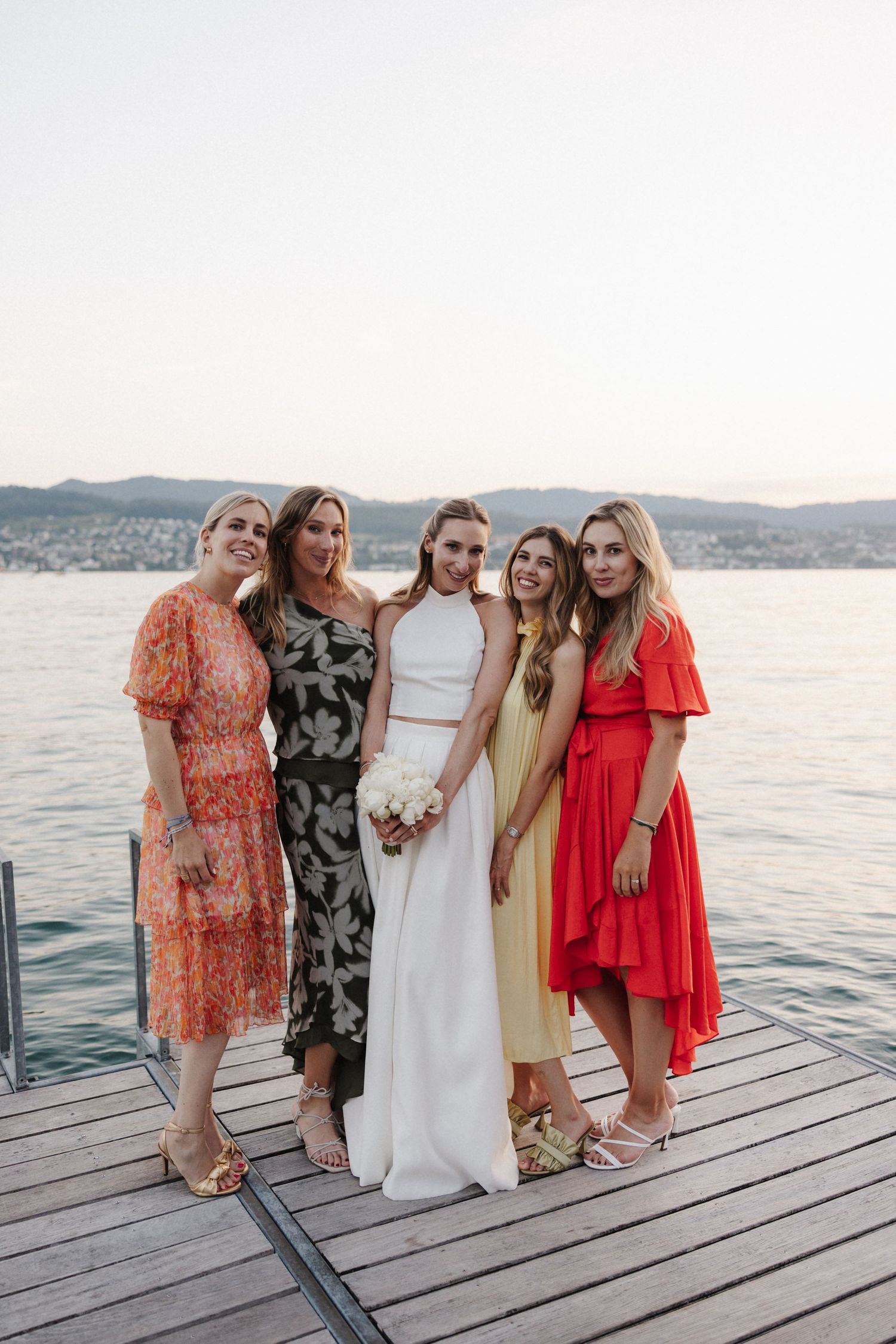 Bride in white holds bouquet posing with four guests in colorful dresses on a lakeside dock.
