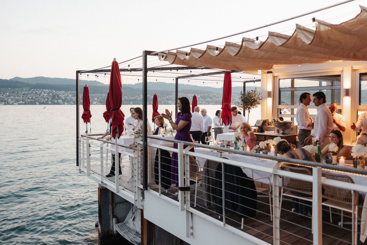 Wedding guests mingle on a stylish waterfront terrace restaurant with lake and mountain views.