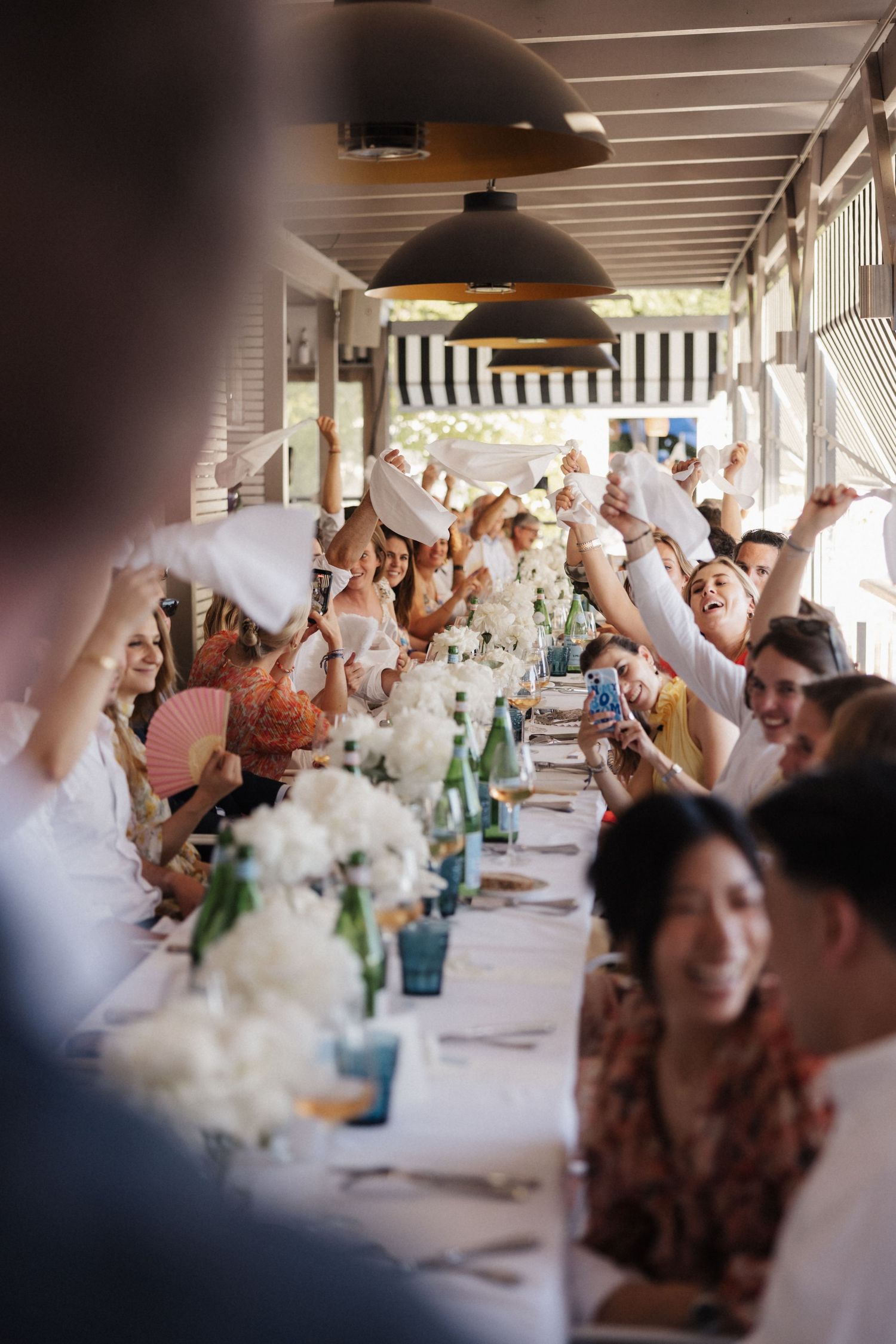 Joyful wedding guests wave white napkins and fans along an elegant reception table with white flower arrangements.