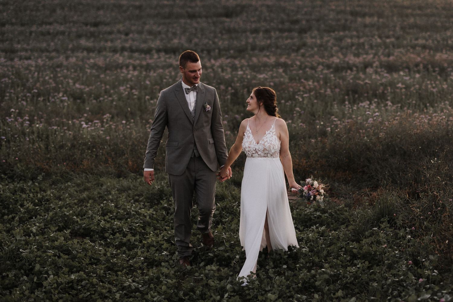 Wedding couple holding hands walking through a blooming field at dusk, bride carrying a bouquet.