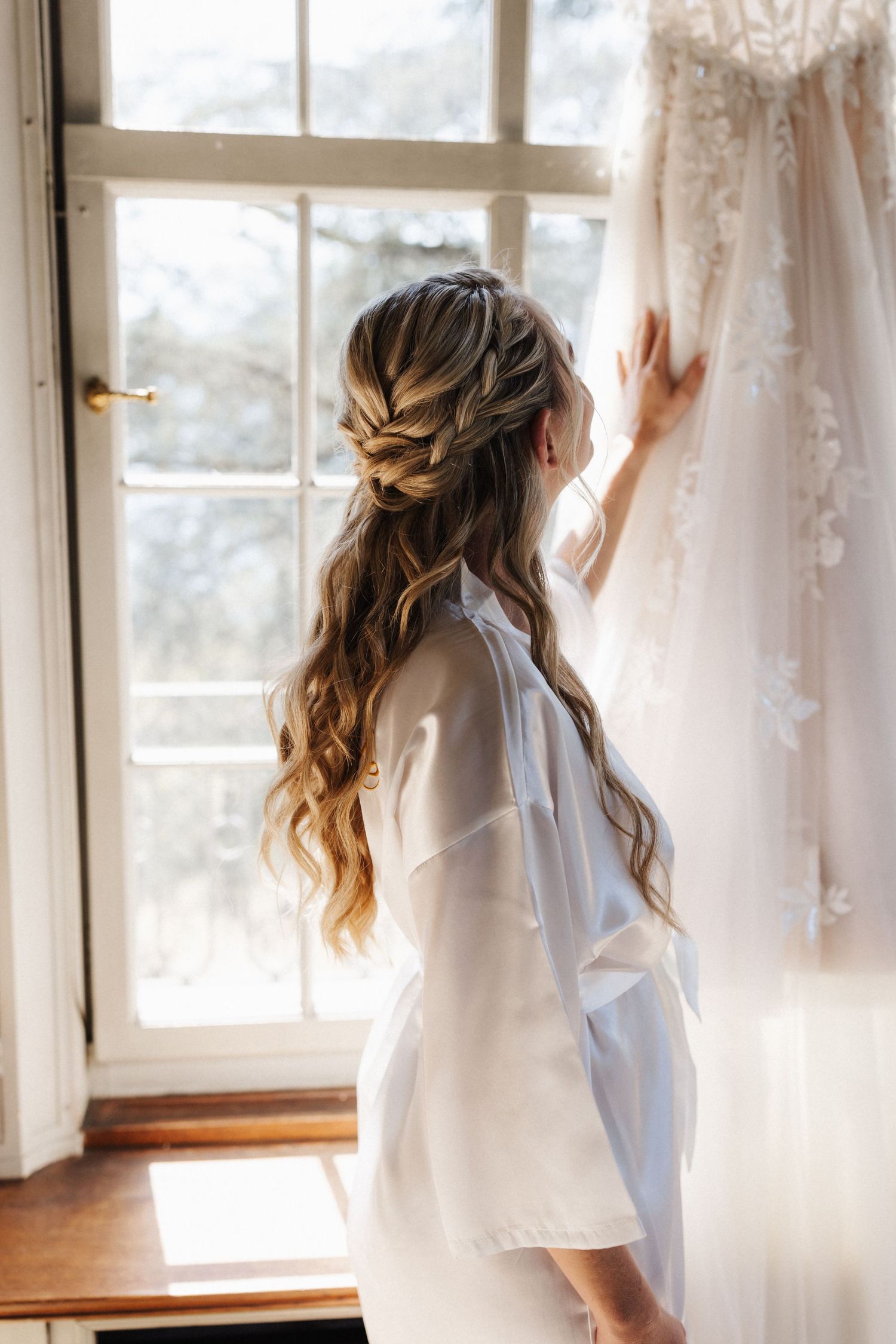 A bride with a braided half-updo hairstyle in a white robe touching her lace wedding gown.