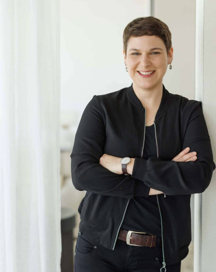 Smiling woman with short hair wearing a black jacket, arms crossed, leaning against a white wall.