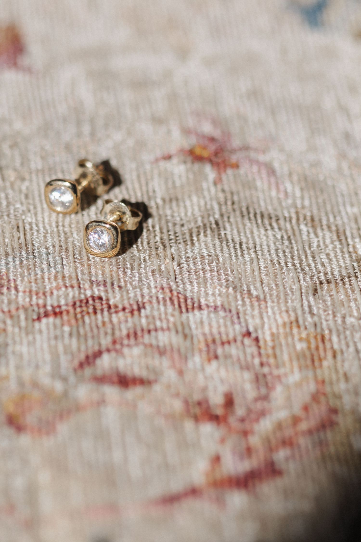 Gold bezel-set diamond stud earrings on a vintage floral rug in sunlight.
