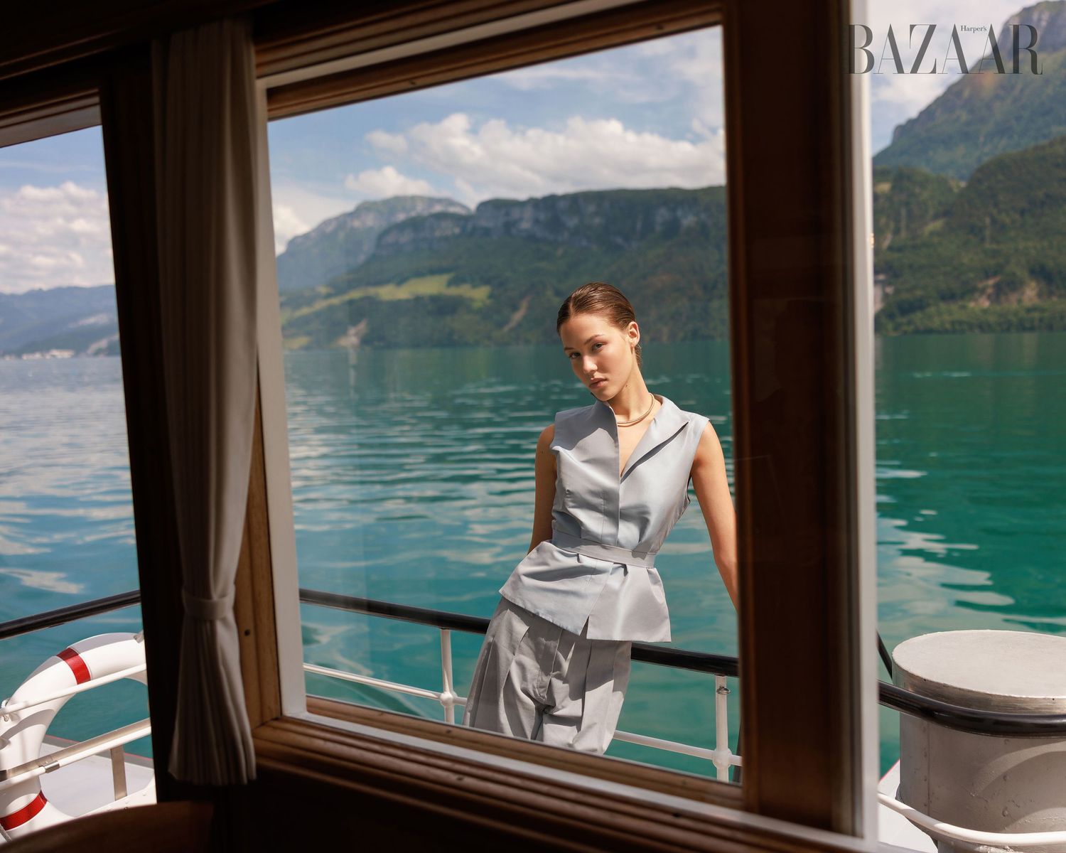 Model in powder blue outfit viewed through boat window with scenic mountain lake backdrop for Harper's Bazaar.