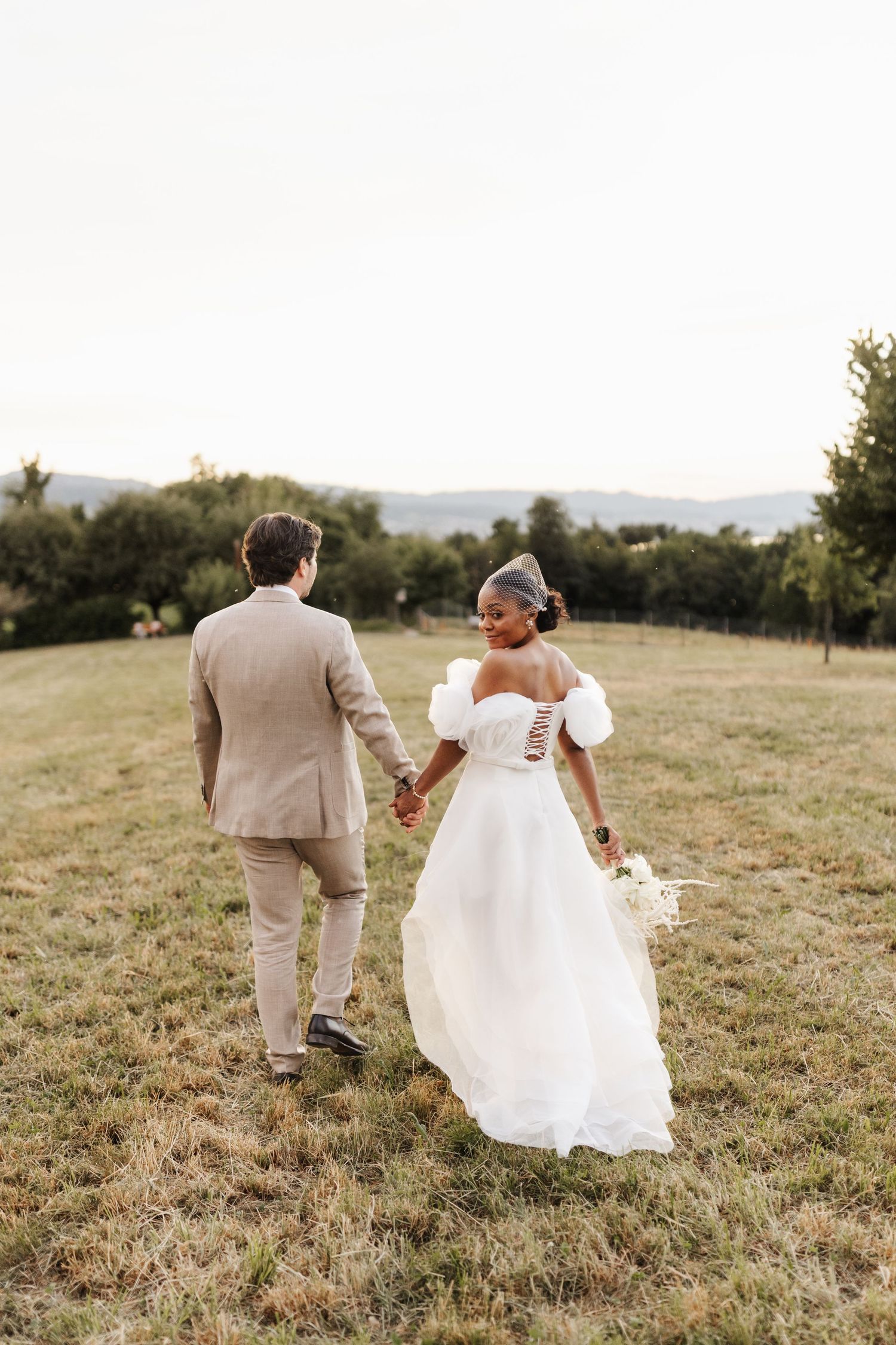 Interracial newlyweds walking hand-in-hand through golden meadow at sunset with mountain backdrop.