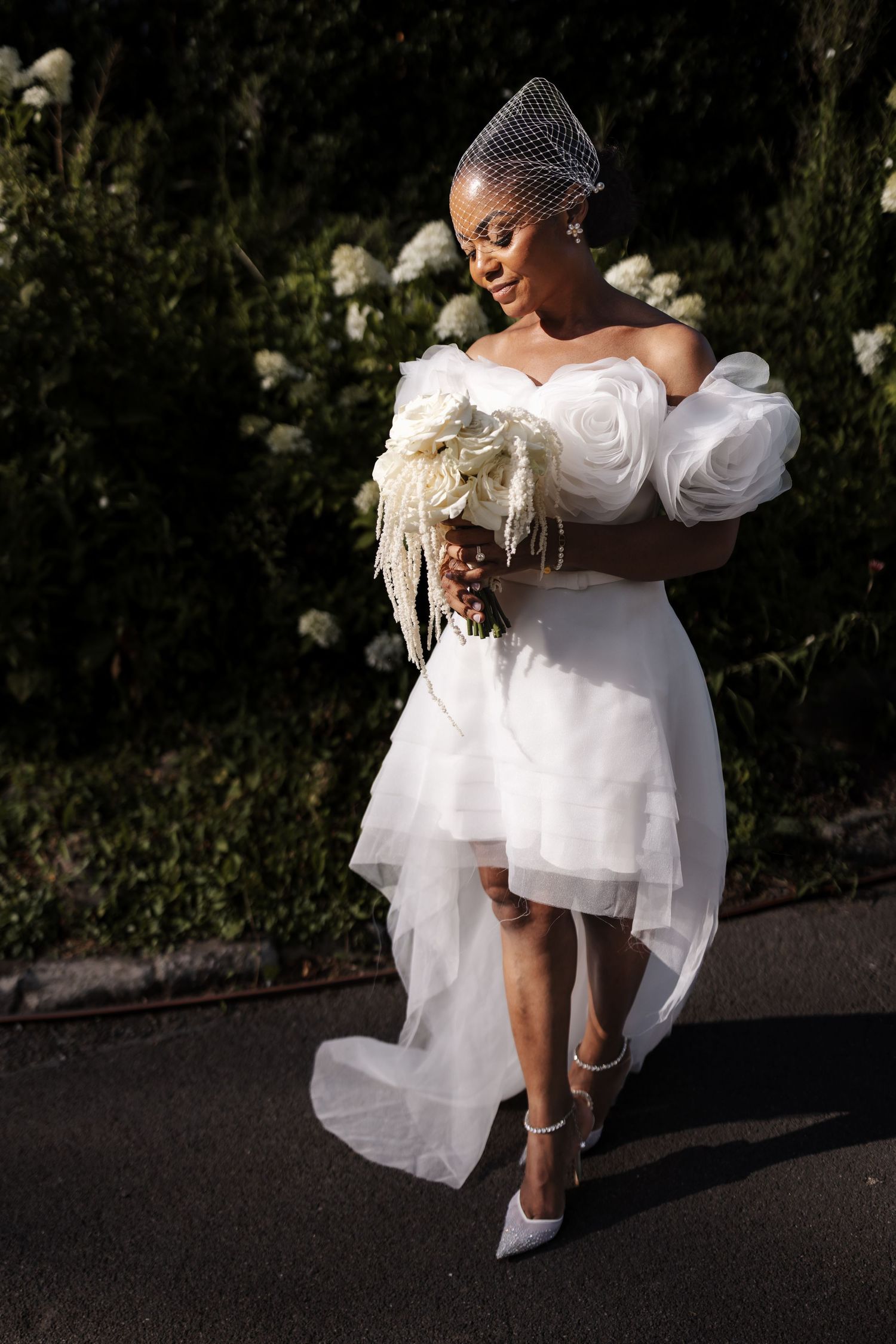 Black bride in white off-shoulder gown with puff sleeves holds floral bouquet outdoors.