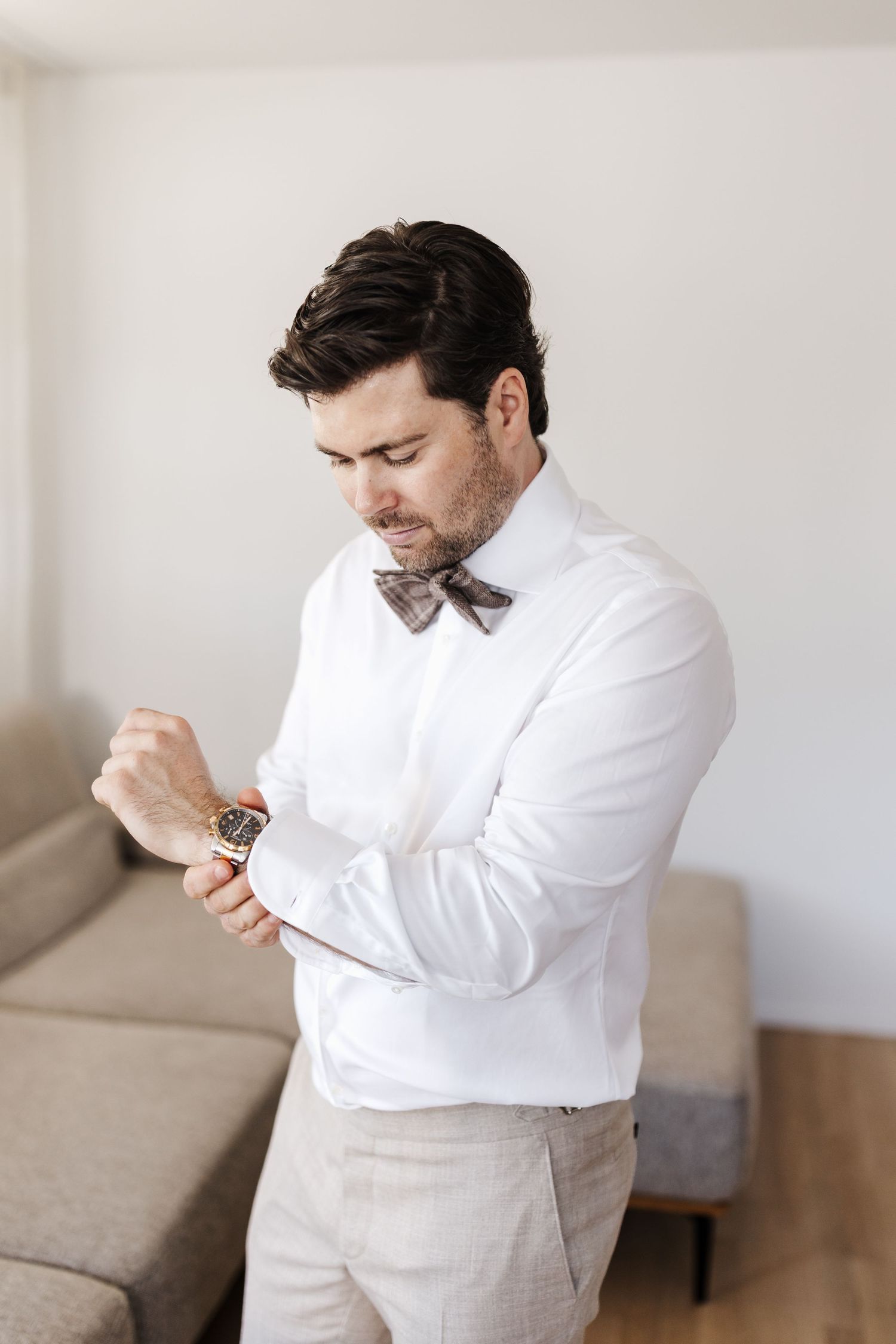 Stylish groom in white shirt and plaid bow tie adjusting his gold watch cufflink indoors.