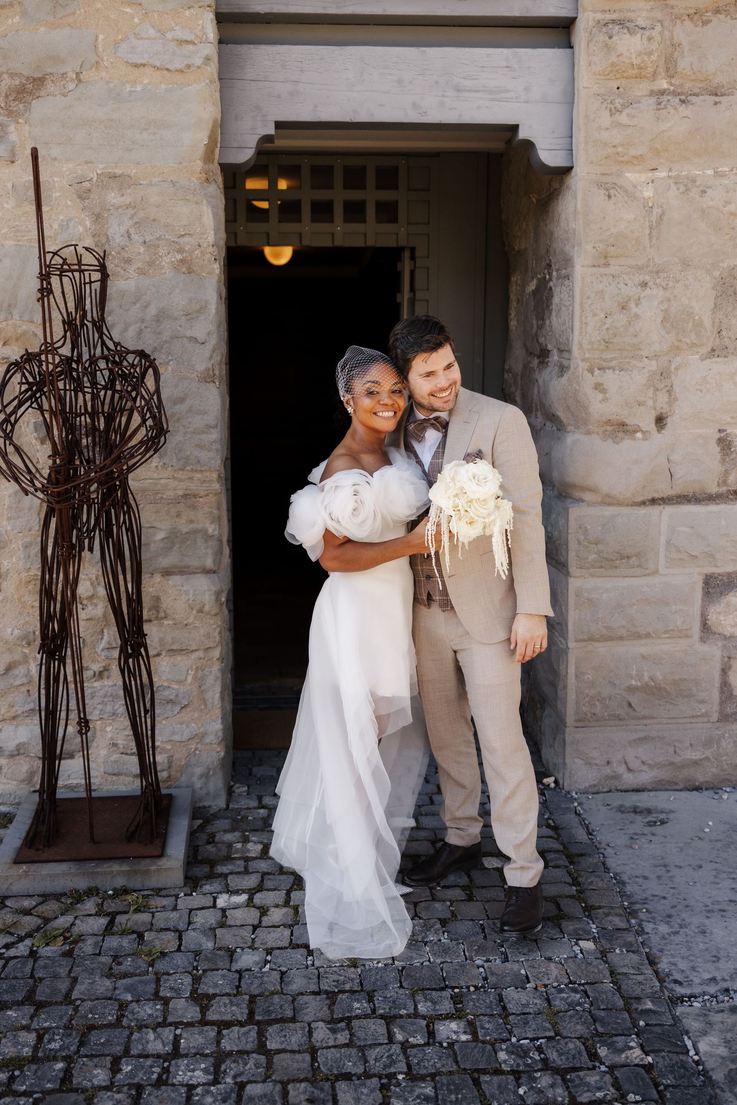 Interracial couple poses in wedding attire outside stone building, bride in white gown holding bouquet, groom in beige suit.