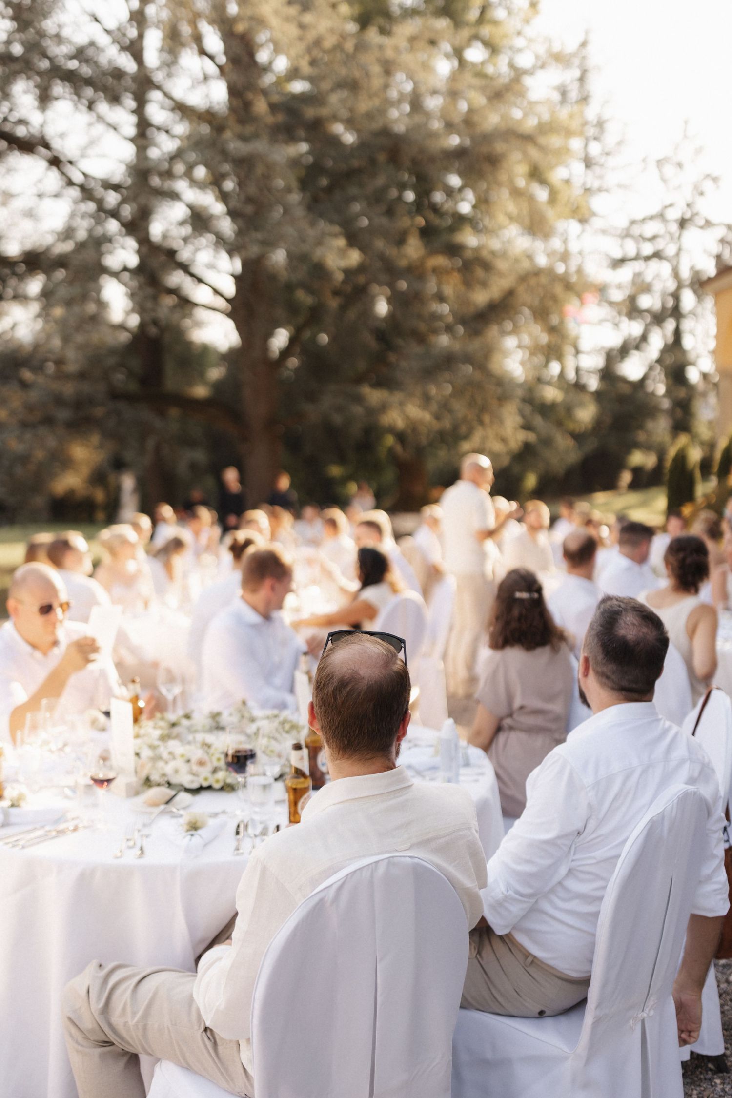 Outdoor wedding reception guests seated at white tables in sunlit garden during golden hour.
