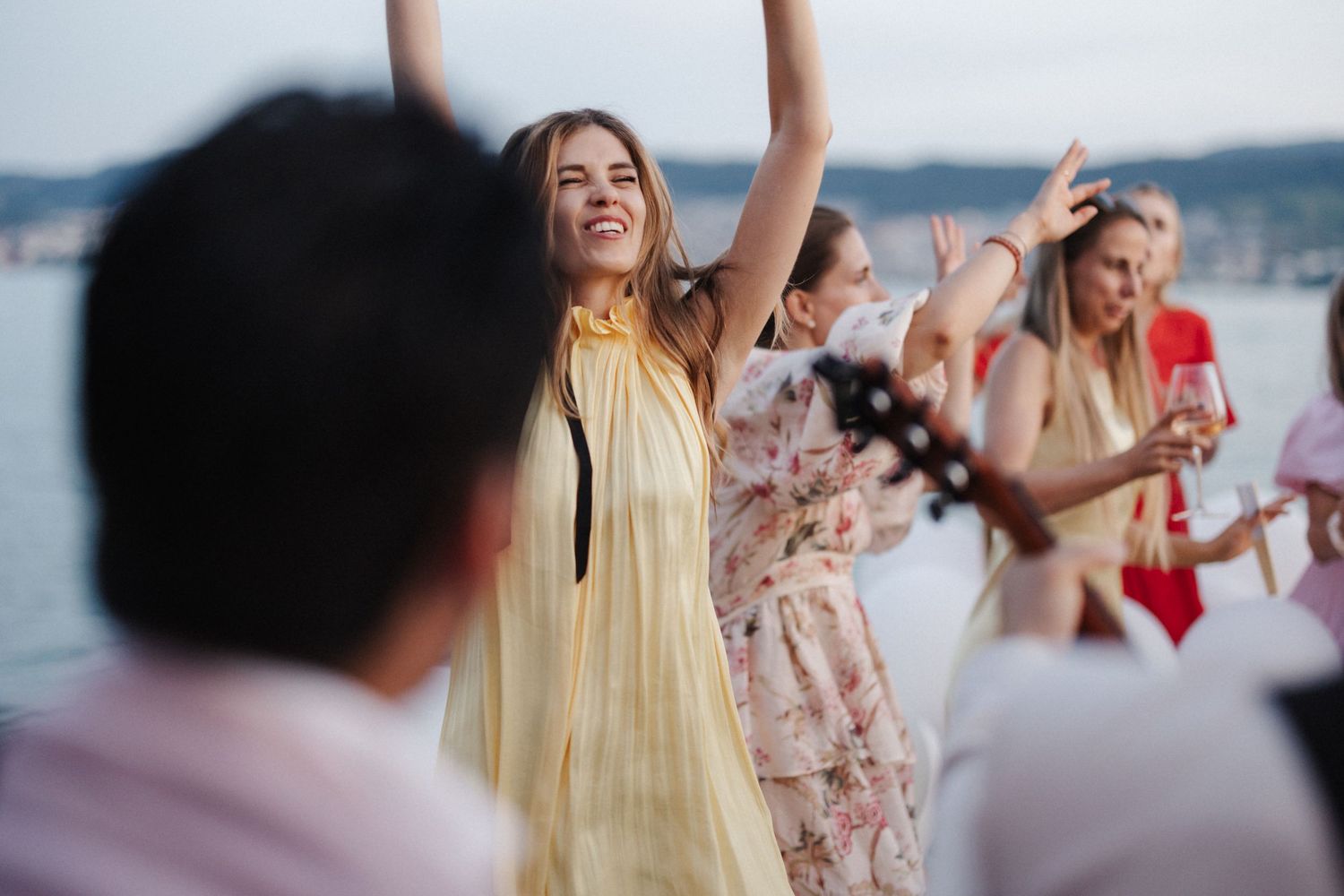 Joyful woman in yellow dress dances with arms raised at an outdoor lakeside wedding celebration.