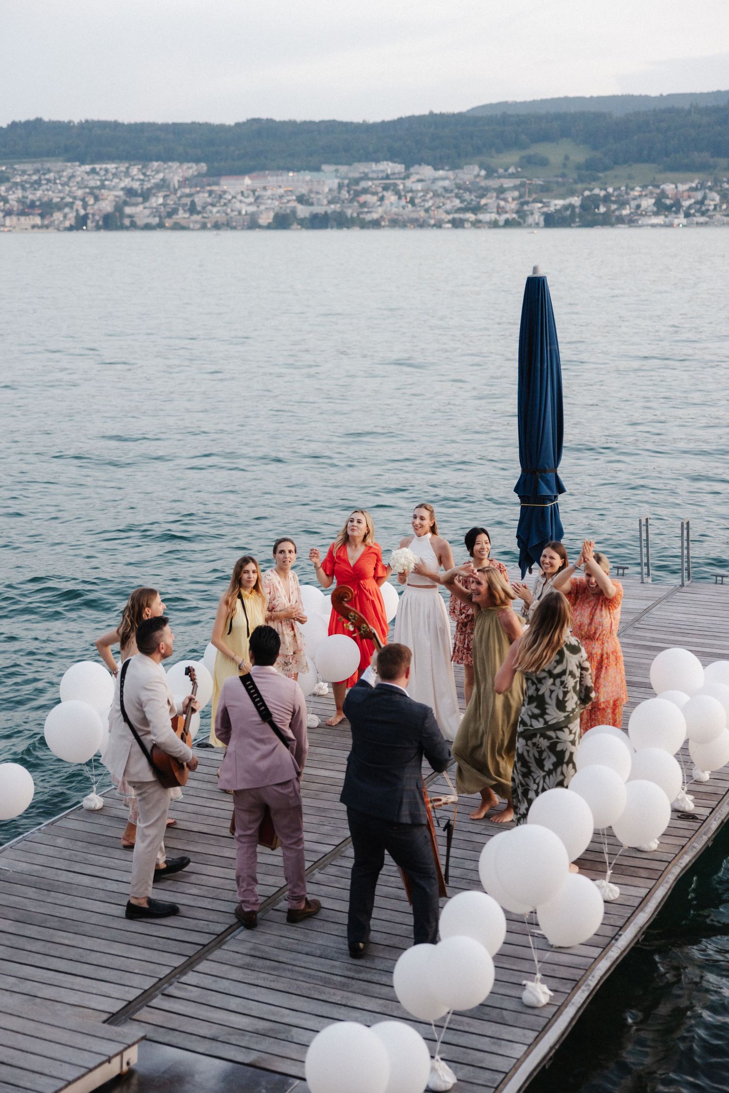Wedding guests gather in a circle on a dock with white balloons while musicians perform lakeside.