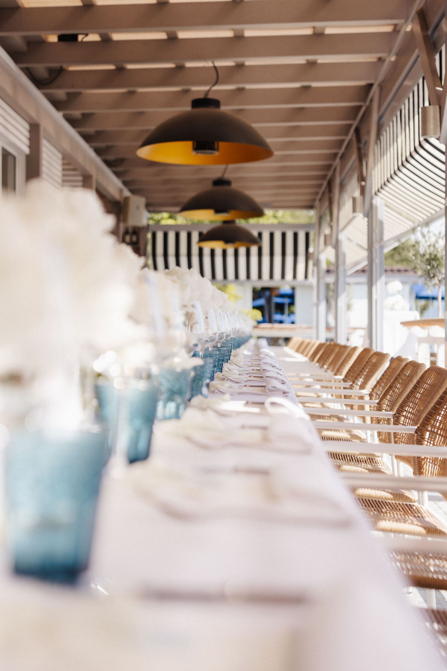 Elegant long dining table set with blue glassware and white floral centerpieces under pendant lights on a covered patio.