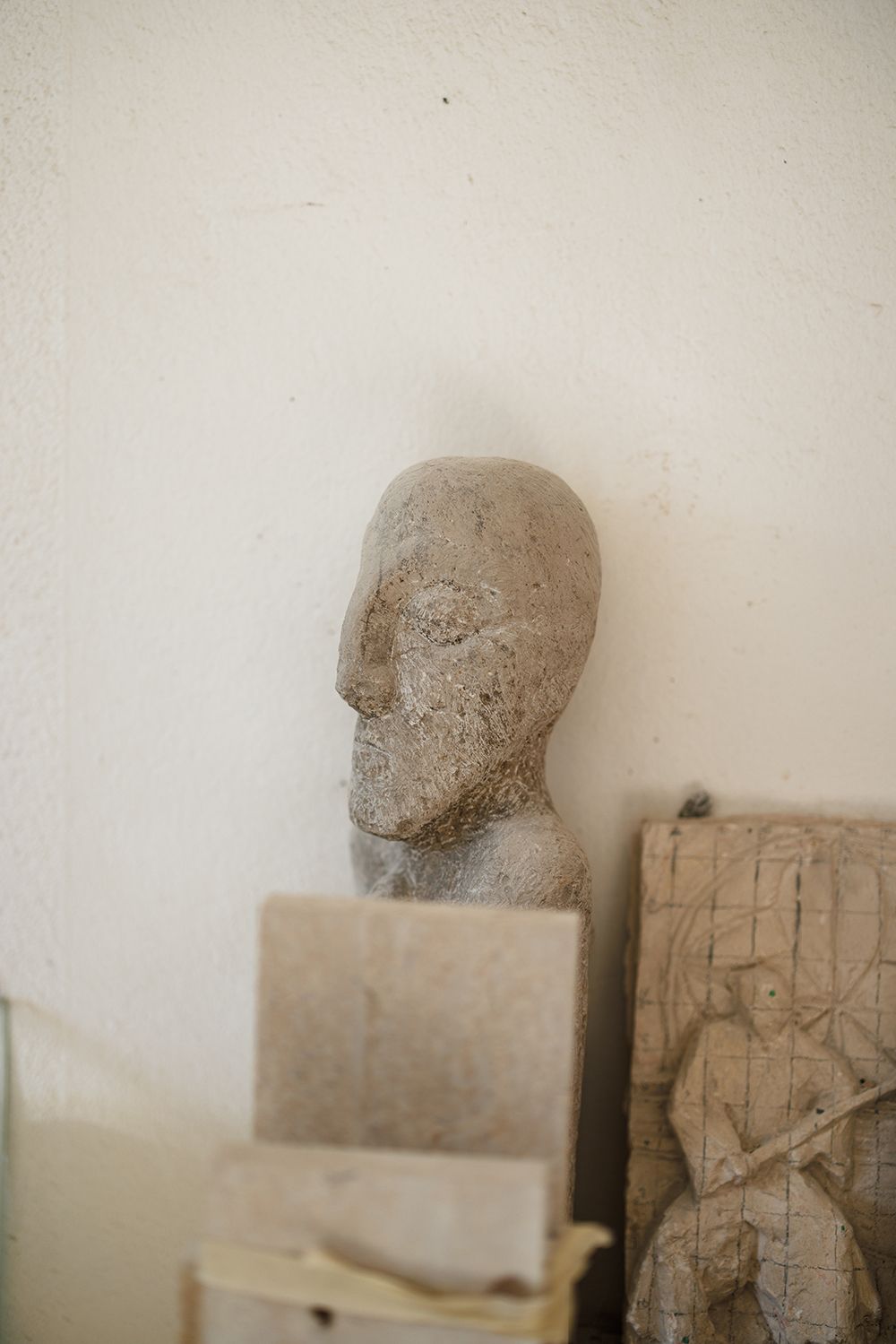 Stone sculpted human head bust displayed against a white wall in an artist's studio with stone slabs nearby.