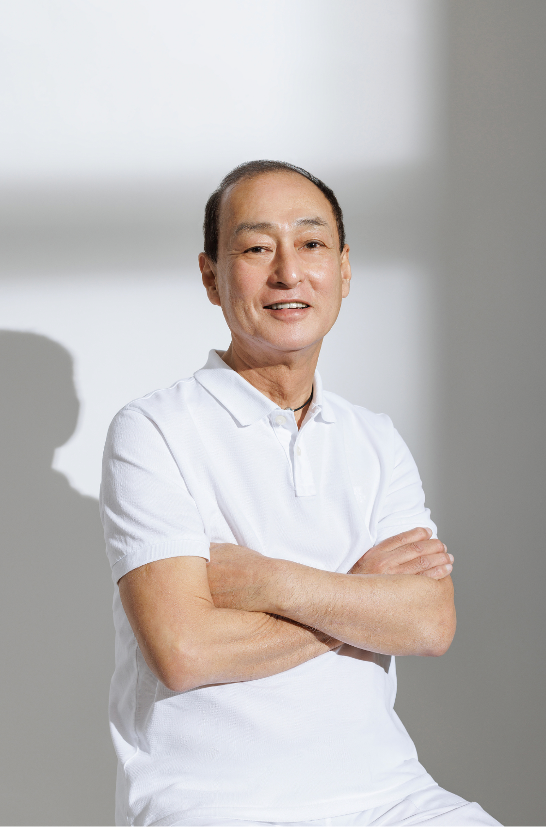 A middle-aged Asian man in a white polo shirt sits with arms crossed, smiling confidently.