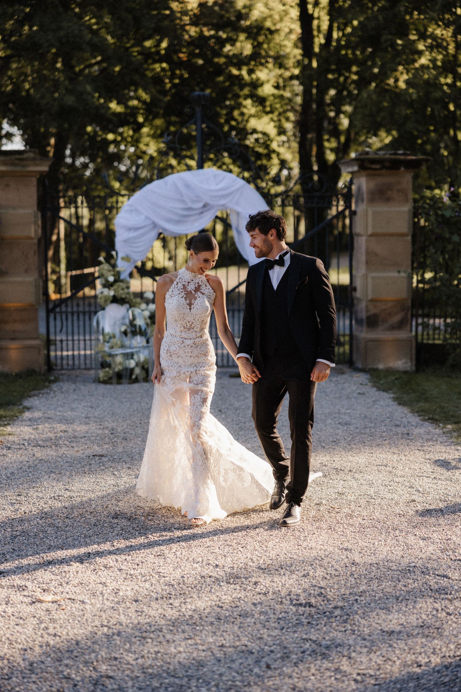 Bride in lace gown and groom in tuxedo walk hand in hand along a gravel path after wedding ceremony.
