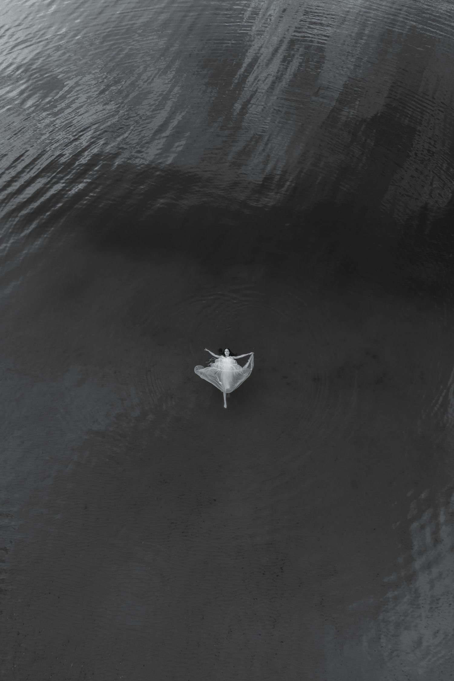 Aerial black and white view of a lone figure in white floating gracefully on a dark rippling water surface.