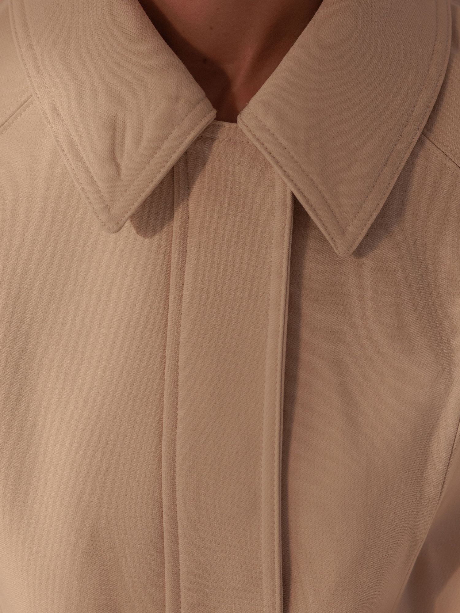 Close-up of a beige collared jacket with clean seam detailing and a hidden button placket.