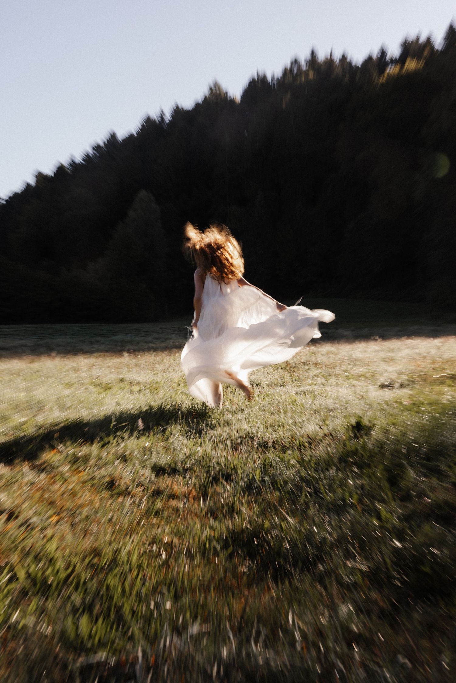 Woman in flowing white dress running through a sunlit meadow with forested mountains in the background.