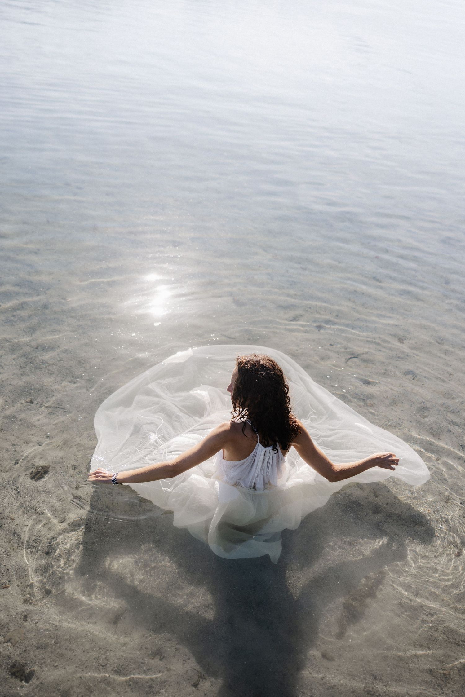 Aerial view of woman in white dress sitting in shallow water with fabric spread around her.