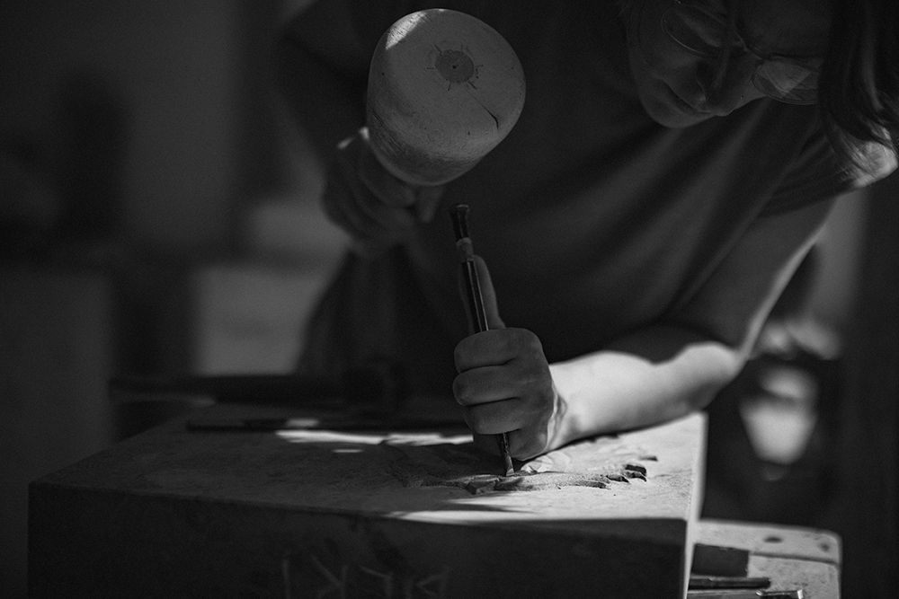 Craftsman uses a mallet and chisel to carve intricate details into a dark wooden surface.