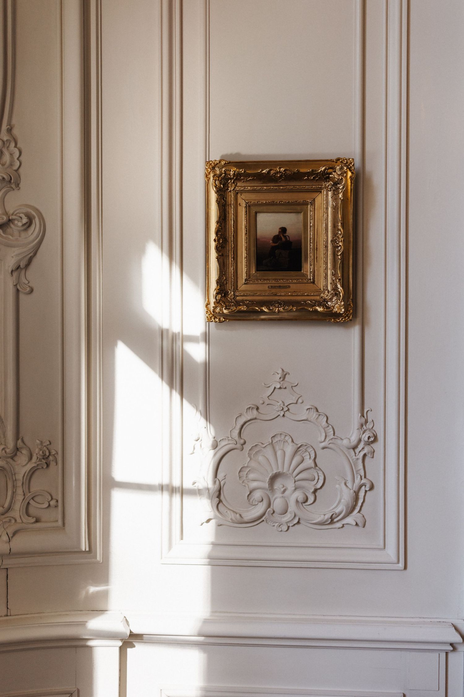Elegant white wall with ornate gold-framed painting and decorative plaster molding in sunlight.