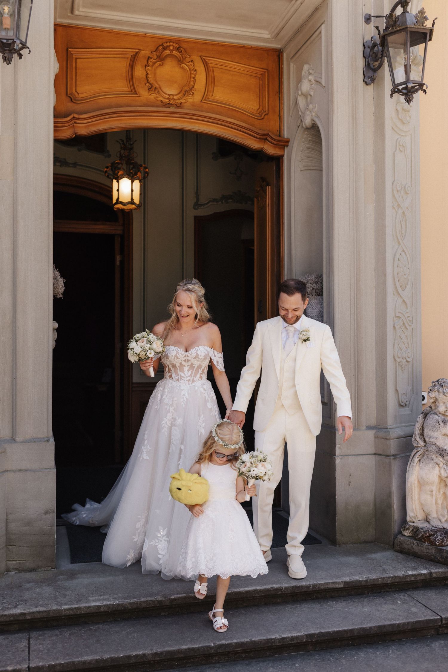 Bride and groom holding hands with flower girl descending ornate church steps on their wedding day.