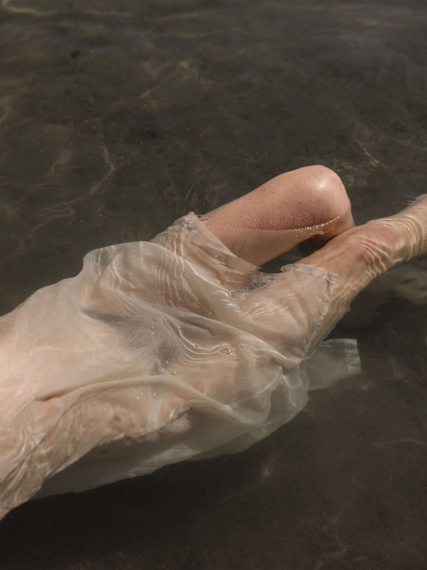 Legs submerged under shallow water covered by sheer white fabric on sandy riverbed.
