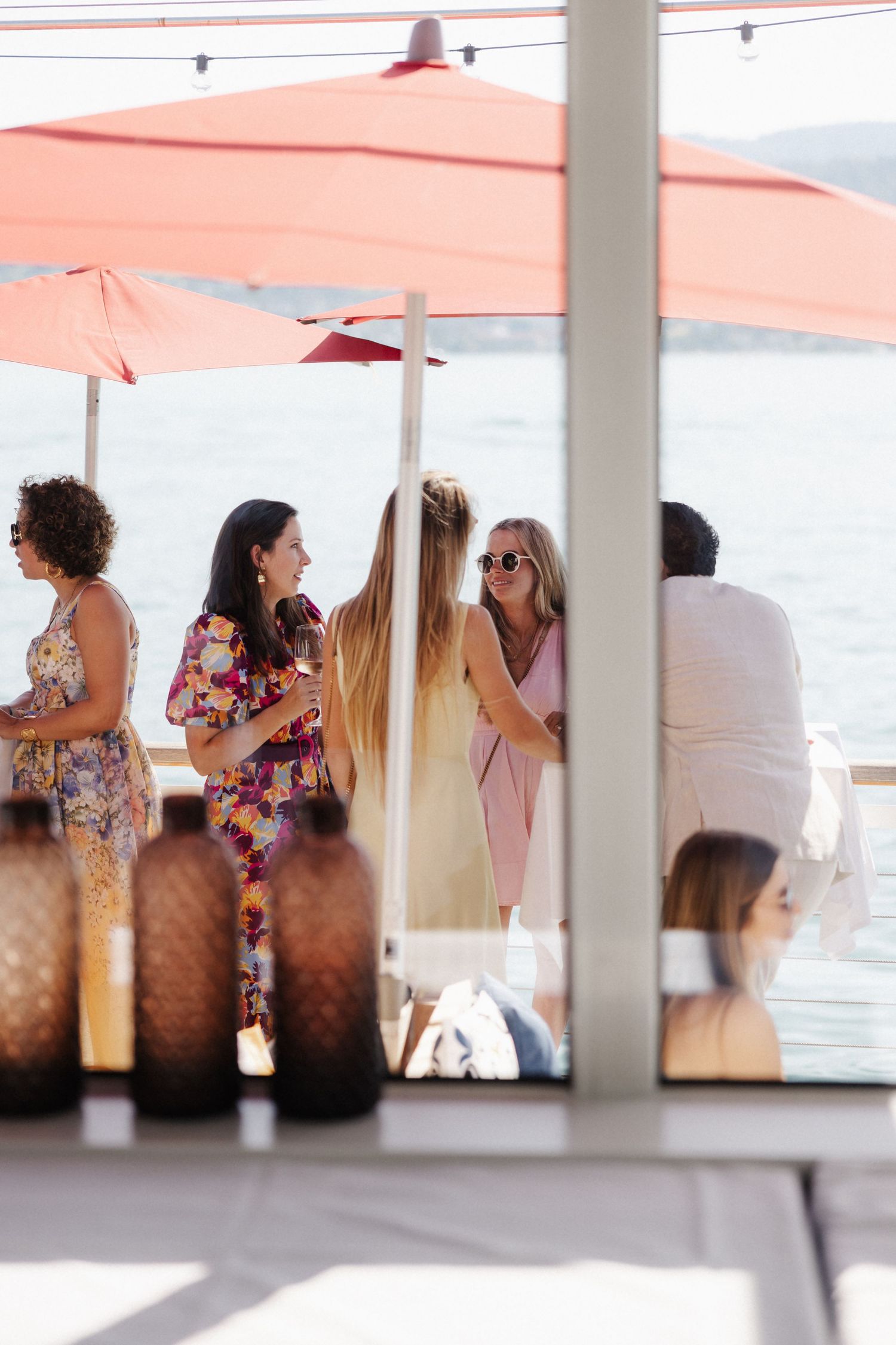 Elegantly dressed wedding guests socializing on a waterfront terrace under red umbrellas in sunlight.