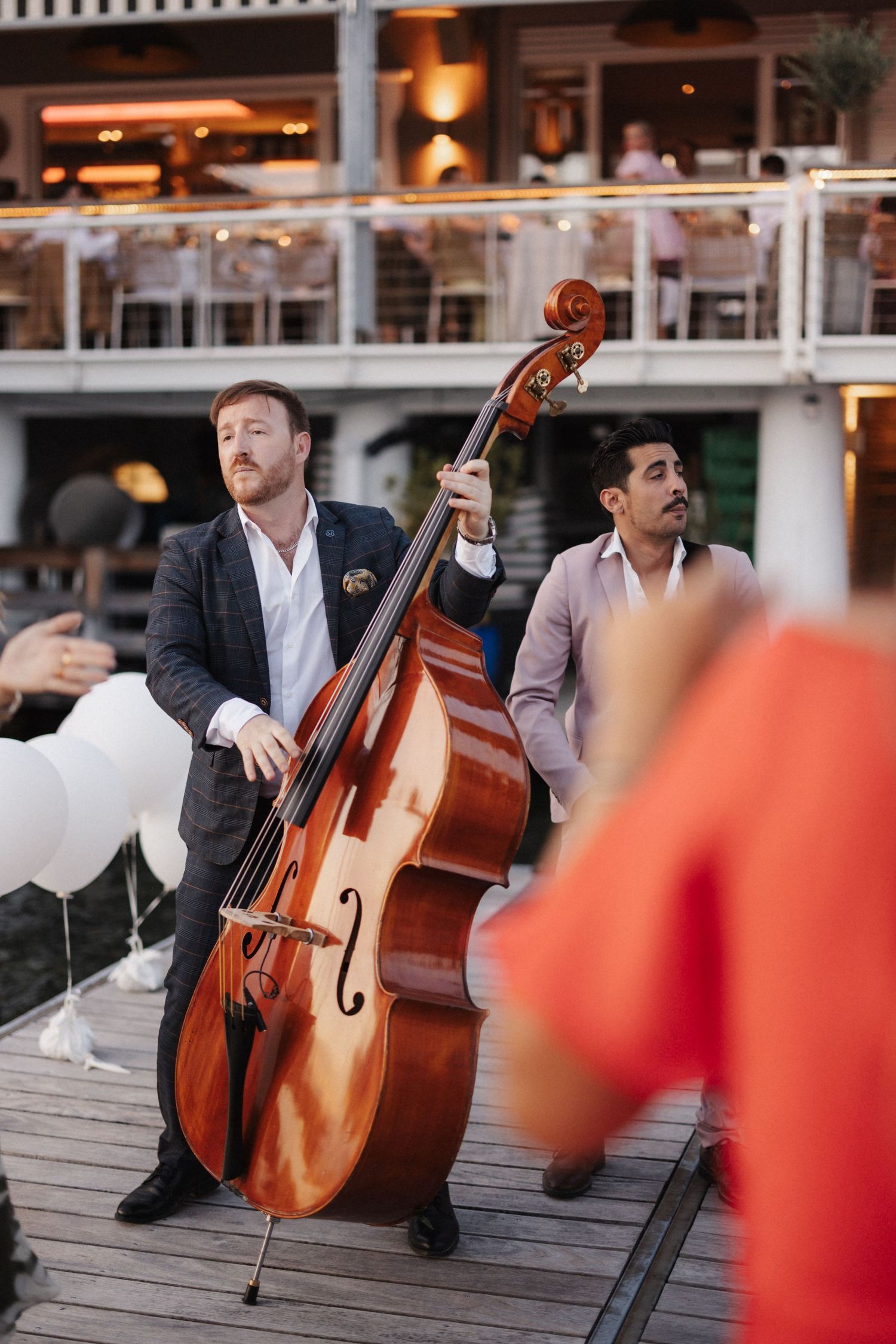 Musician in dark suit plays double bass on a waterfront dock during an outdoor wedding celebration.