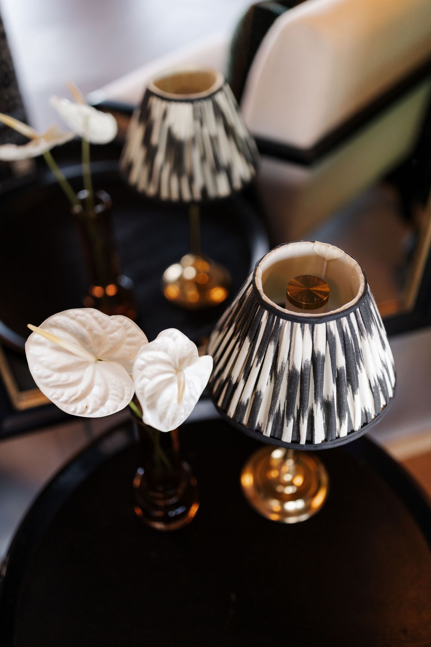 Black decorative table with white anthurium flowers and two black and gold ikat patterned lamp shades.