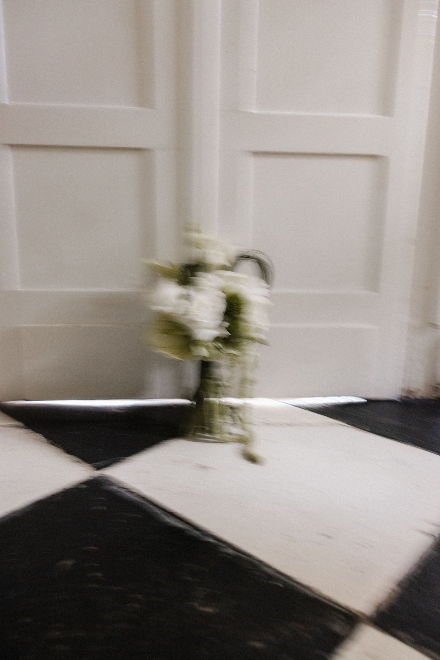 White wedding bouquet in vase placed against white paneled door on checkered tile floor.