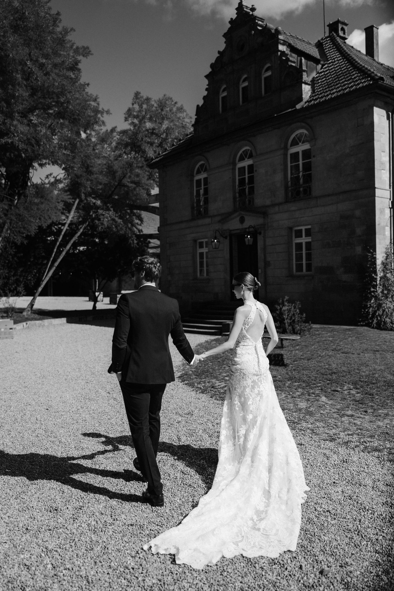 Black and white photo of bride and groom walking hand in hand toward a historic stone manor.
