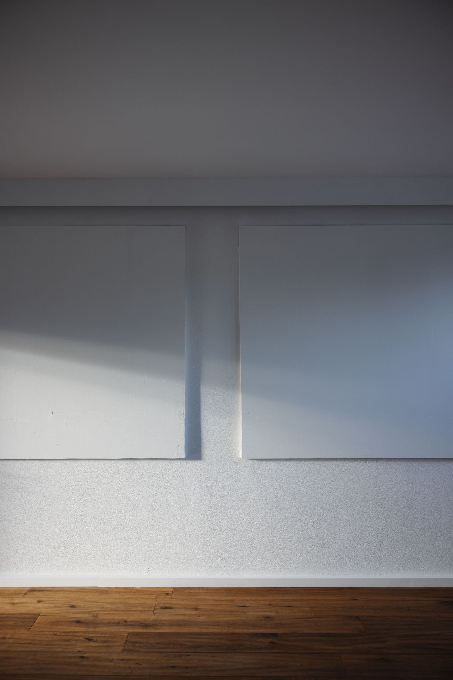 Two large white canvases on white wall with natural light shadows and wooden floor.