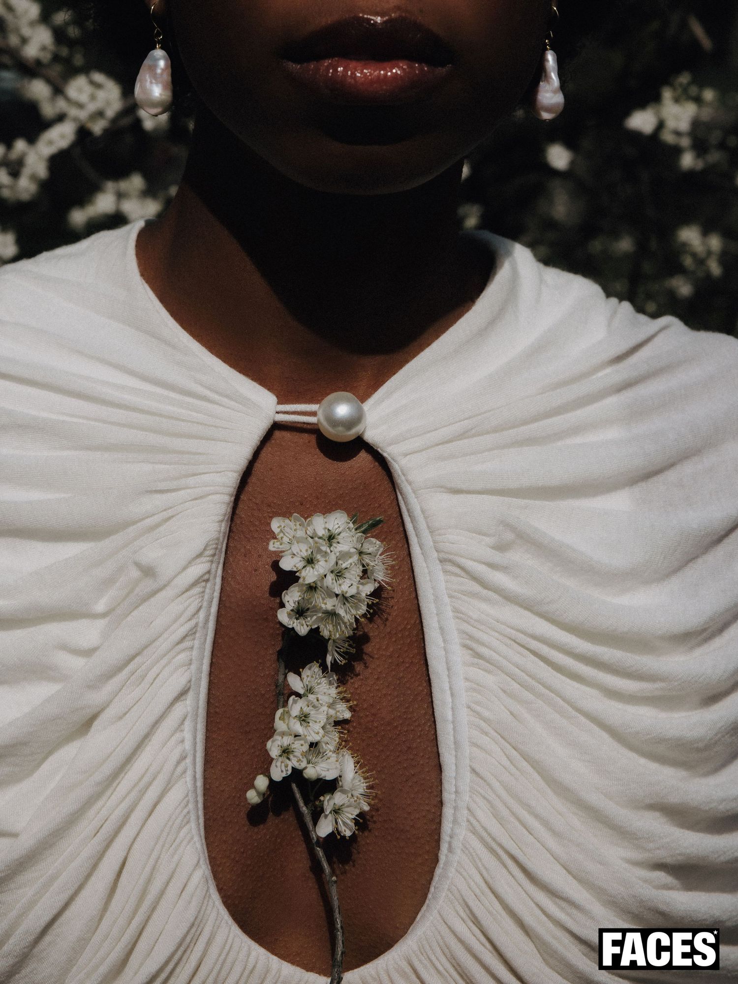A Black woman wears pearl earrings and a white draped dress adorned with a cherry blossom branch brooch.